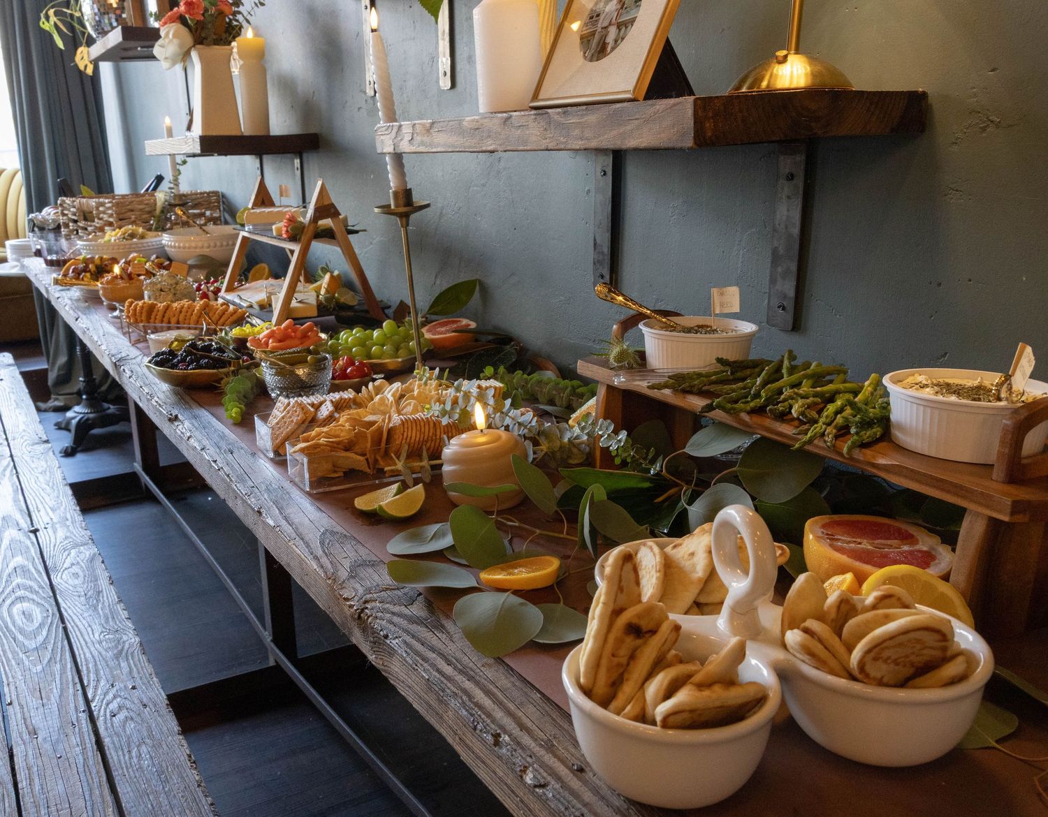 A rustic wooden table displays a variety of fresh baked breads and pastries in baskets under warm pendant lighting.