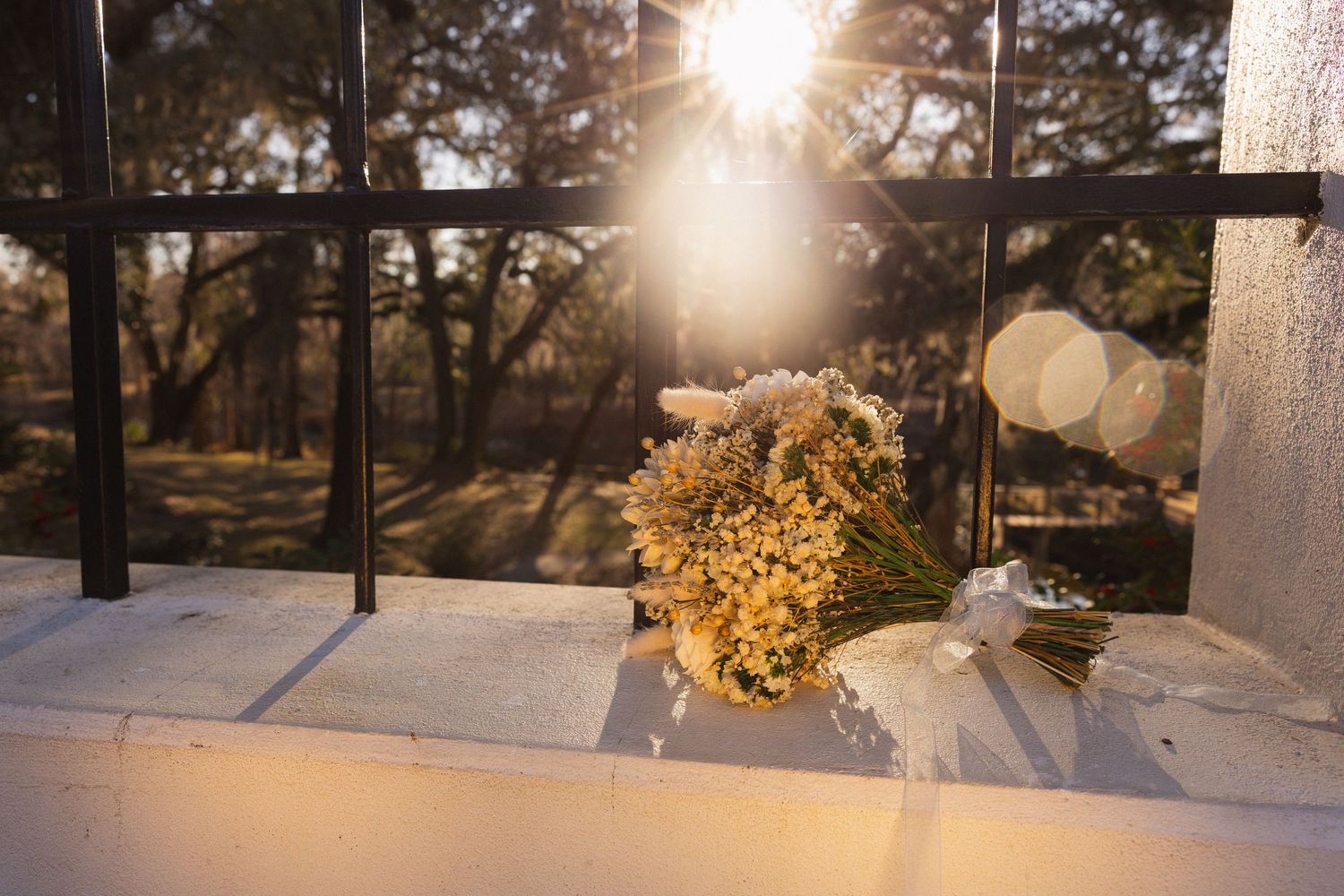 A sunlit wedding bouquet with yellow flowers rests on a white surface with sunbeams streaming through trees in the background.
