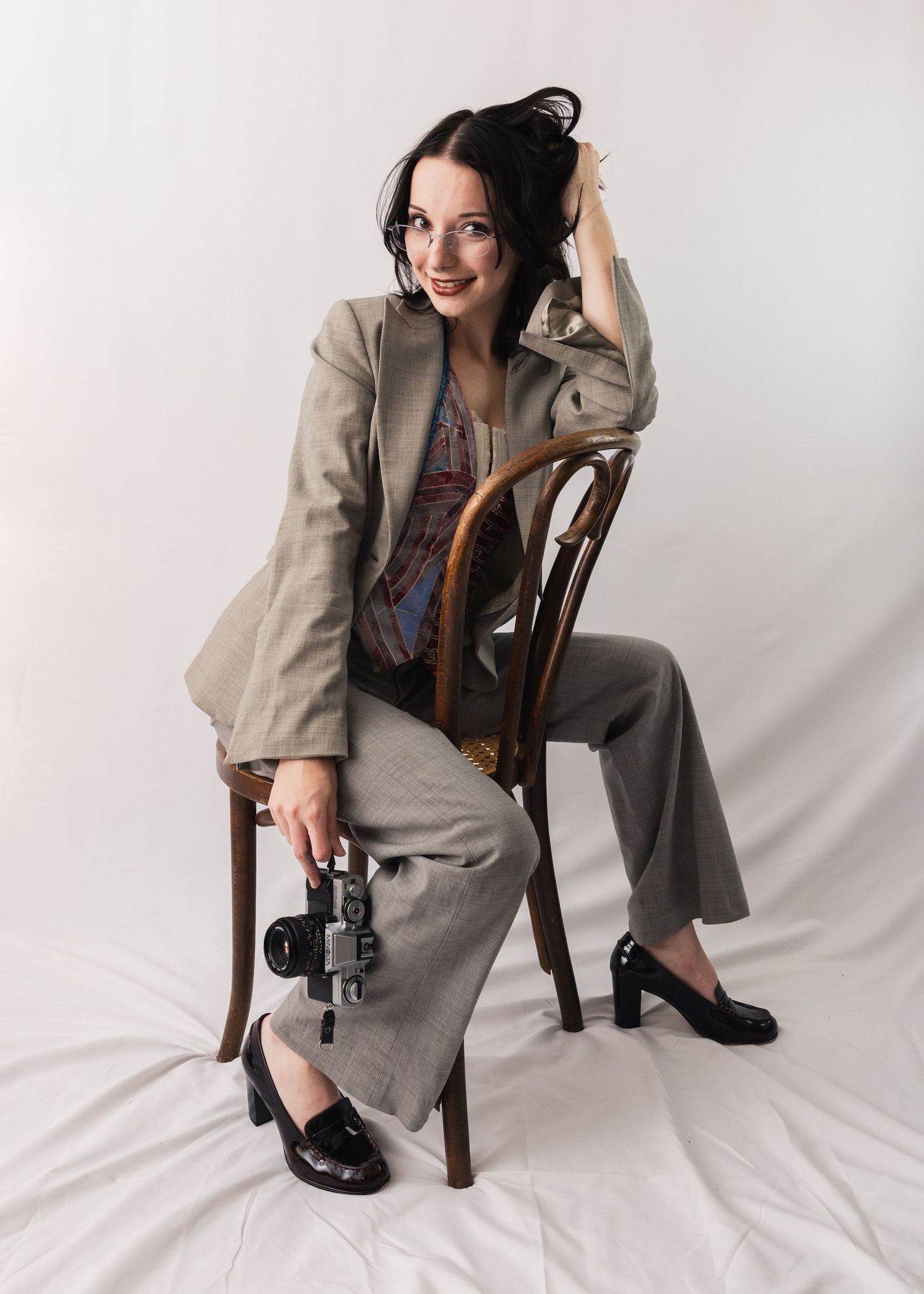 Woman in gray suit holding camera seated on wooden chair, smiling against white backdrop.