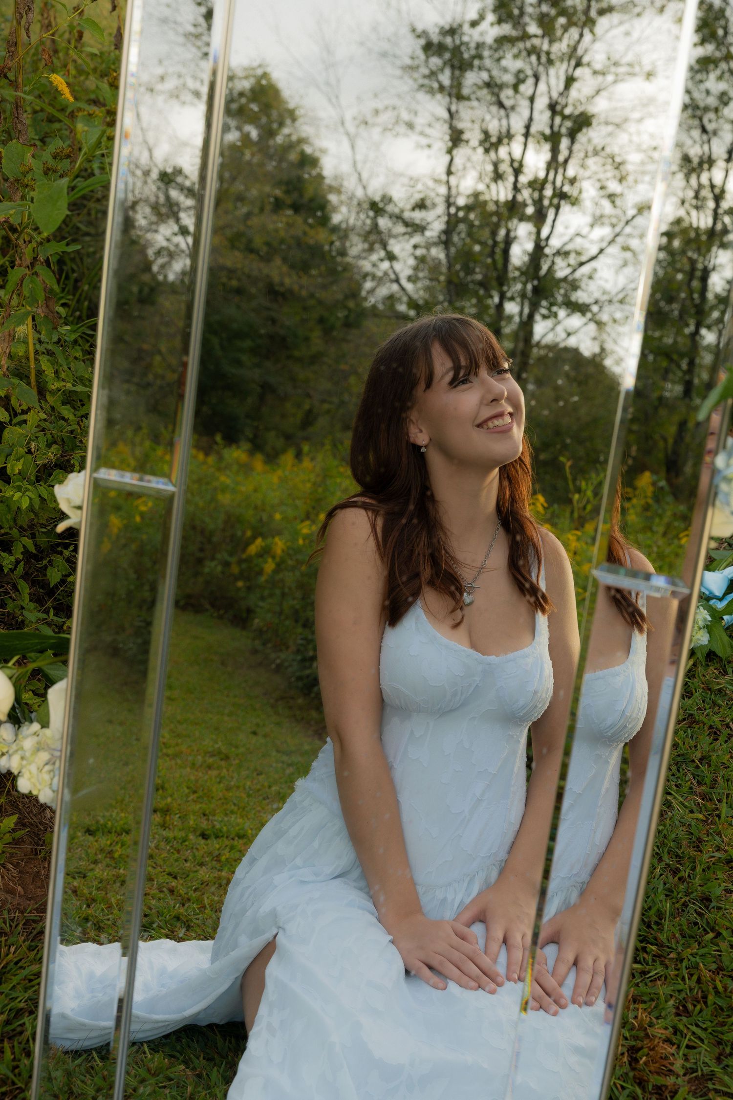 A person in a light blue dress sits on a mirrored surface outdoors surrounded by greenery and trees.