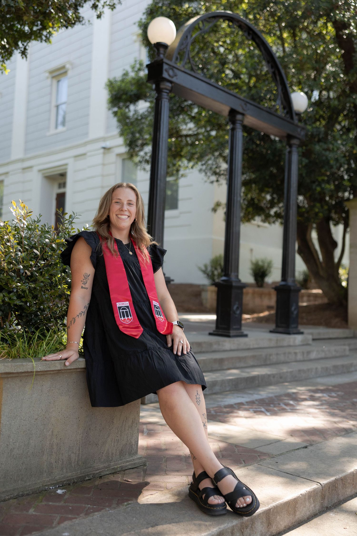 University of Georgia Graduate in black dress sitting on stone wall near iconic campus archway.