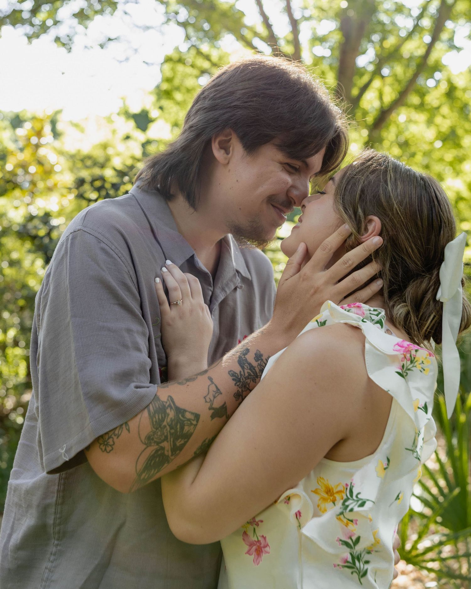 Two people share a tender romantic moment outdoors in a garden setting with sunlit trees in the background.