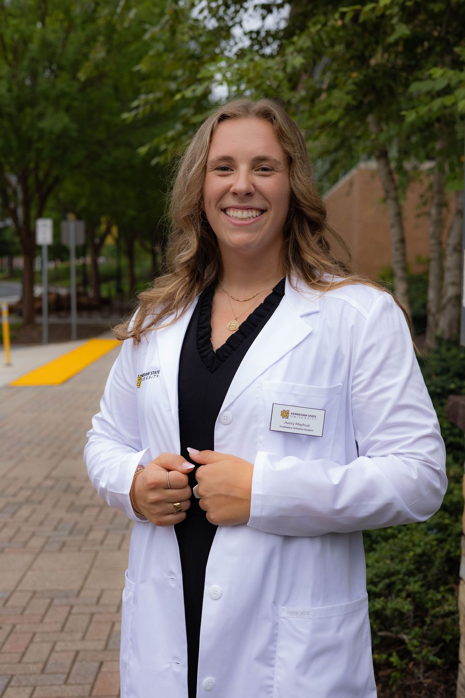 Healthcare professional in white lab coat and black shirt standing on brick pathway with trees in background.
