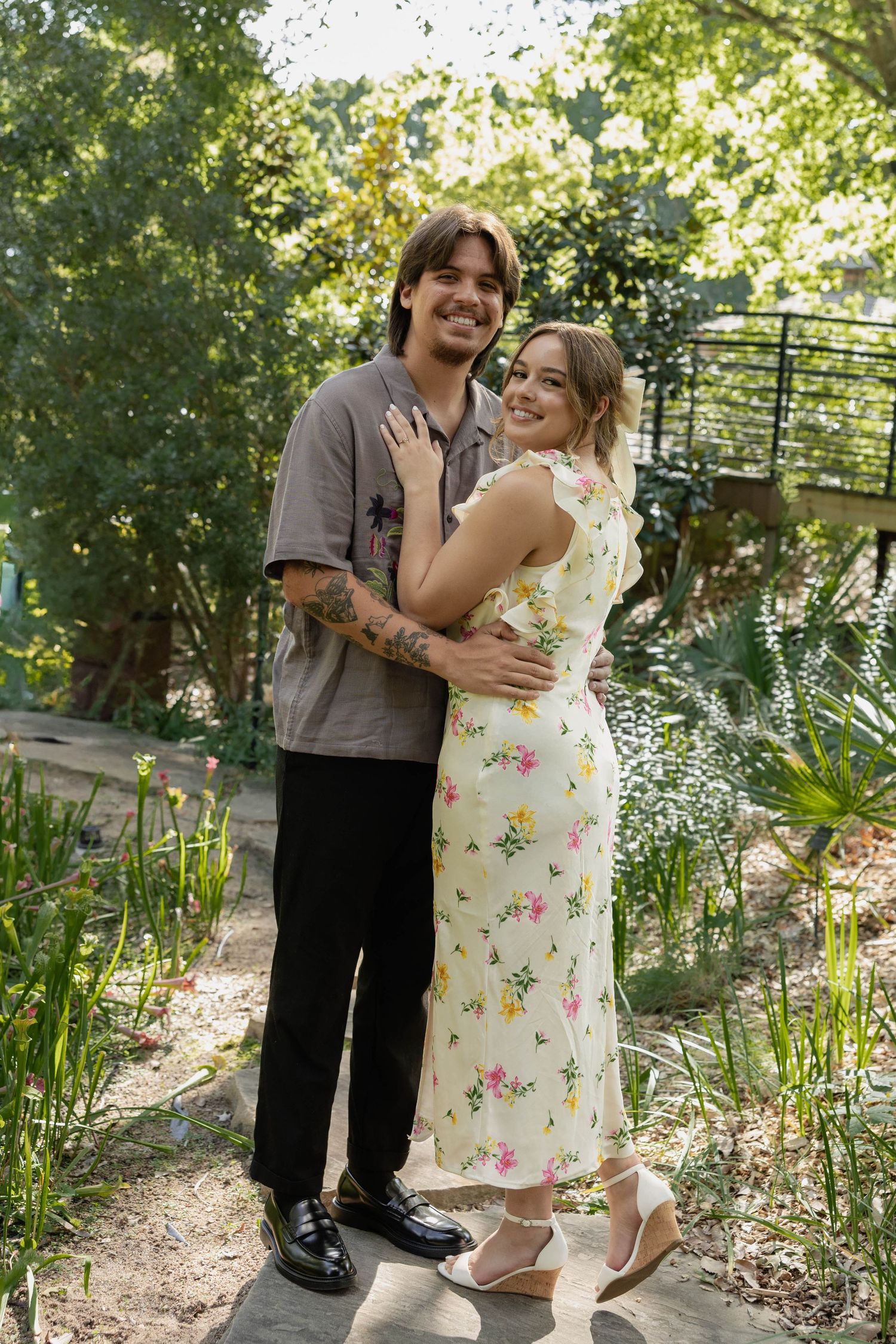 A loving couple embraces in a sunlit garden pathway surrounded by lush greenery on a warm summer day.