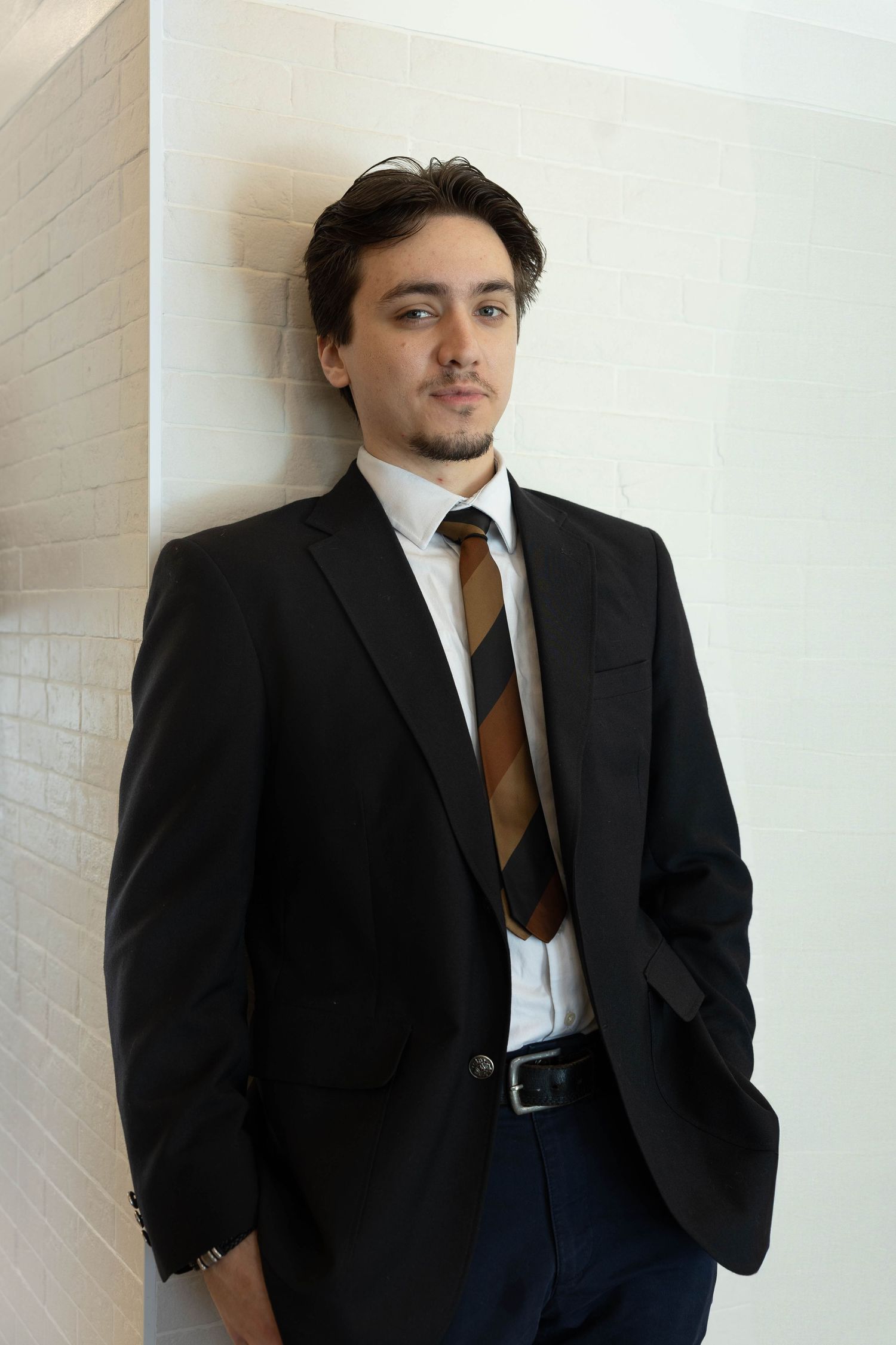 A person in a black suit and brown tie poses against a white brick wall in a professional portrait series.