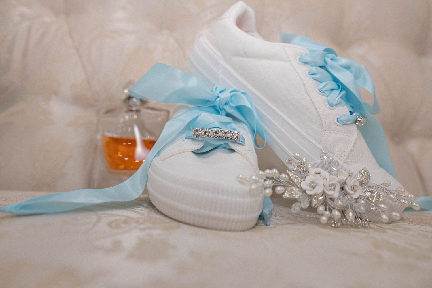 White baby shoes with blue ribbons and delicate pearl accessories lay on a soft cream fabric background.
