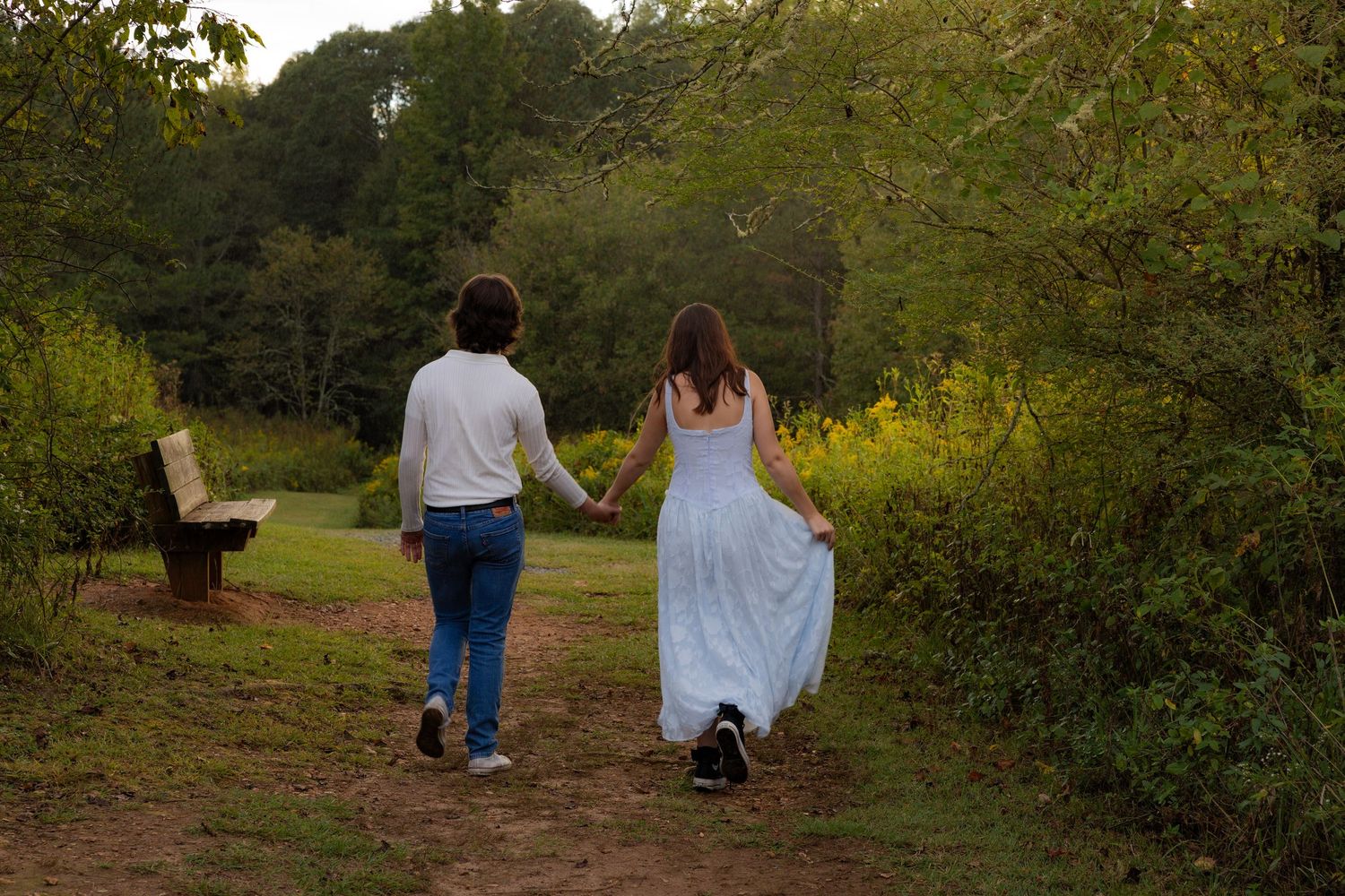 A couple walks hand in hand through a lush green park pathway during a romantic outdoor photoshoot.