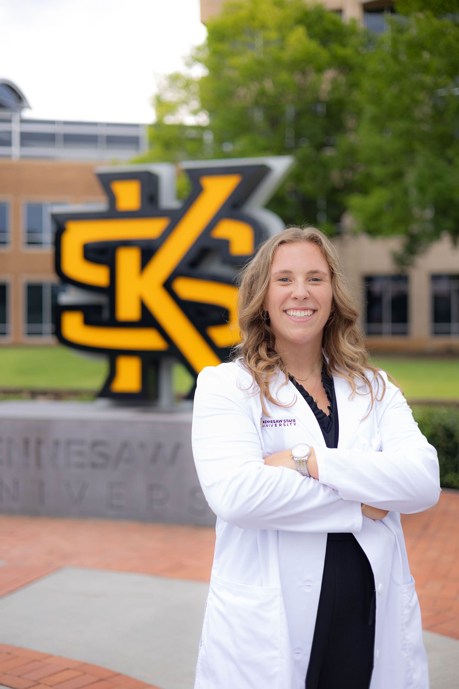 A medical professional in a white coat stands confidently on a university campus with Kennesaw State University signage visible.