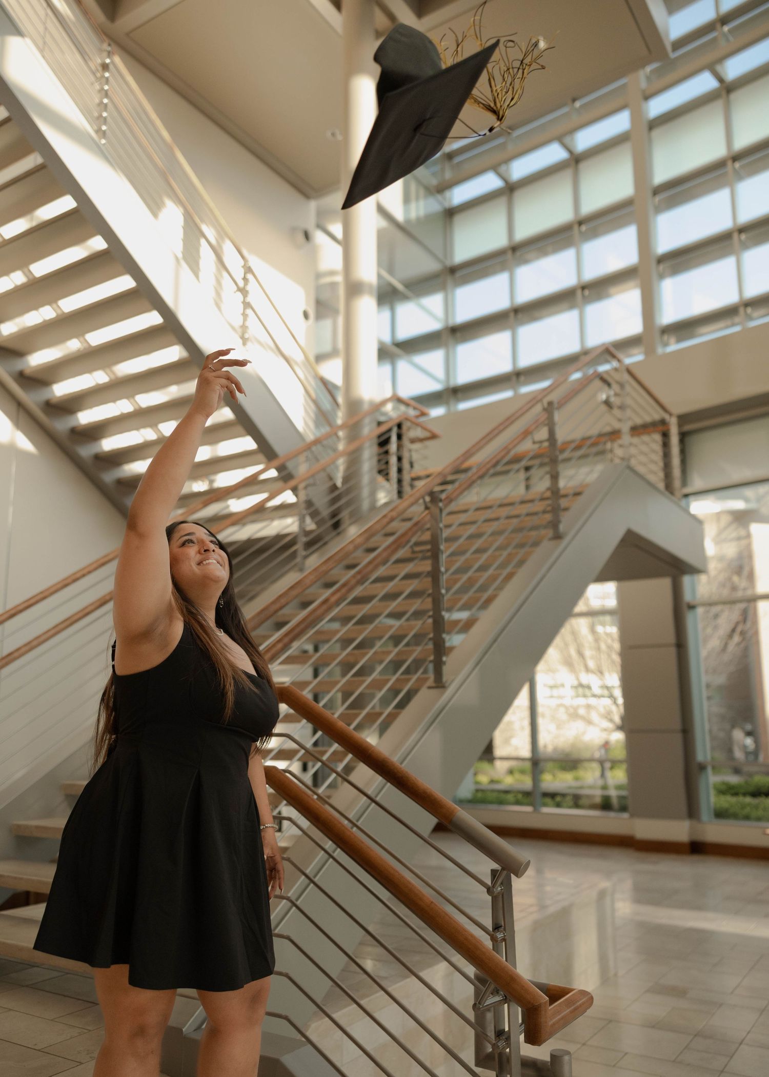 A graduate tosses their black mortarboard cap in celebration inside a modern building with glass windows and stairs.