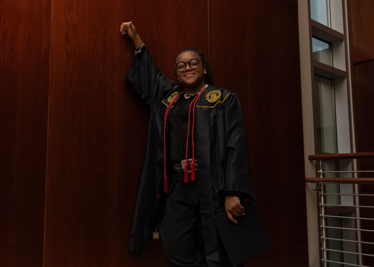 A person stands in a dark room with wooden paneling wearing a black hoodie and glasses.