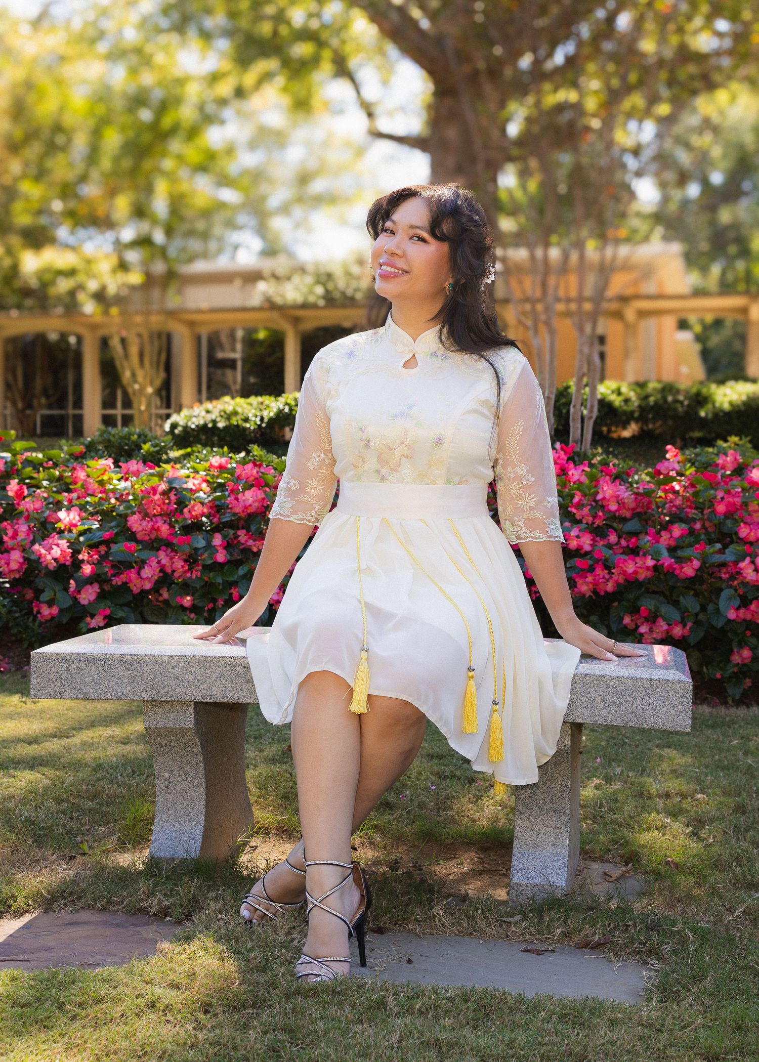 A woman in a white lace dress sits gracefully on a stone bench surrounded by vibrant pink rose bushes in a garden setting.