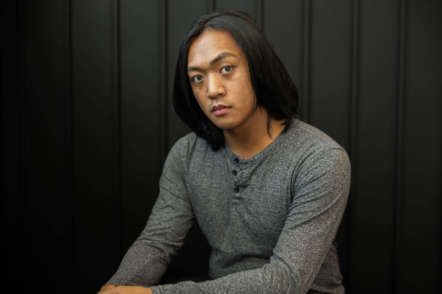 Portrait photograph of a person with long straight black hair wearing a gray henley shirt against a dark background.