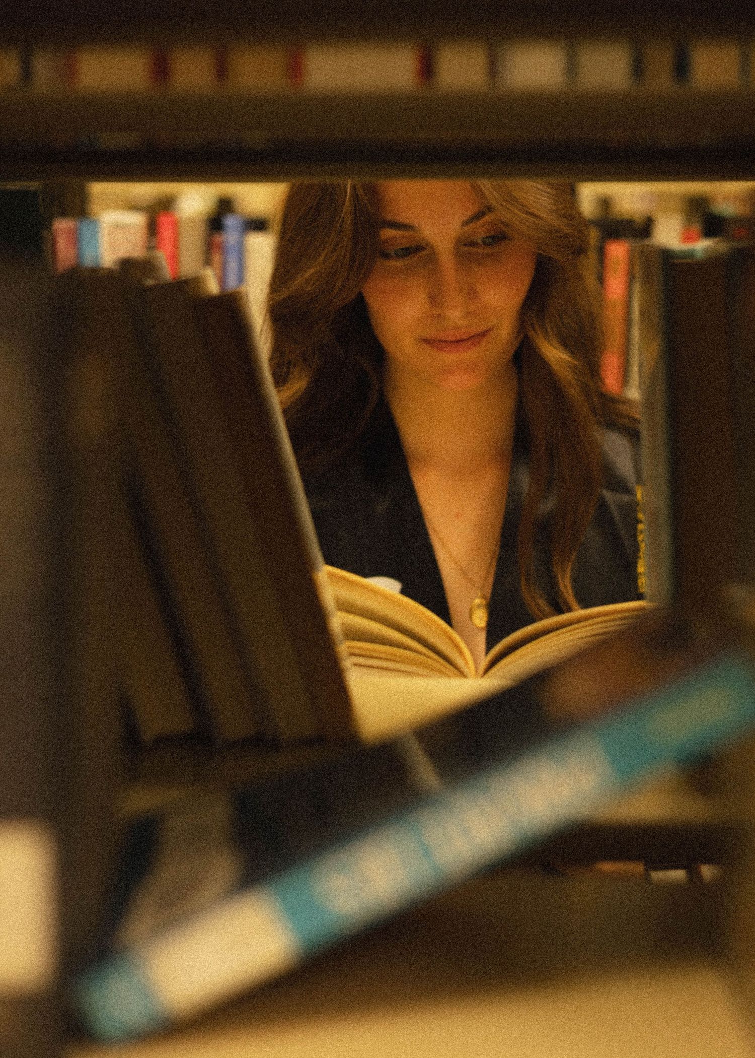 Student reading an open book between library stacks in warm lighting.