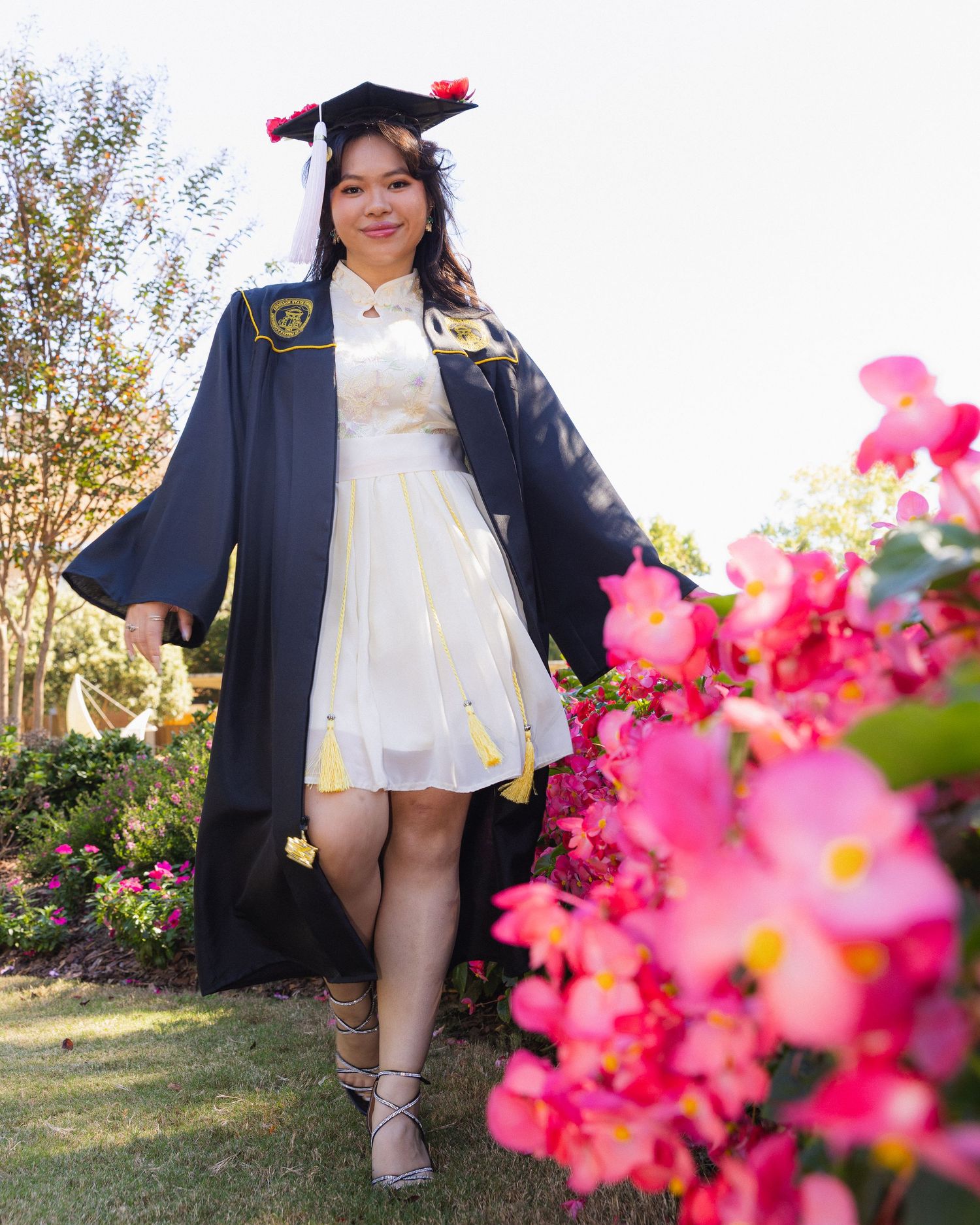 Graduate in cap and gown poses next to vibrant pink bougainvillea flowers in a garden setting during commencement.
