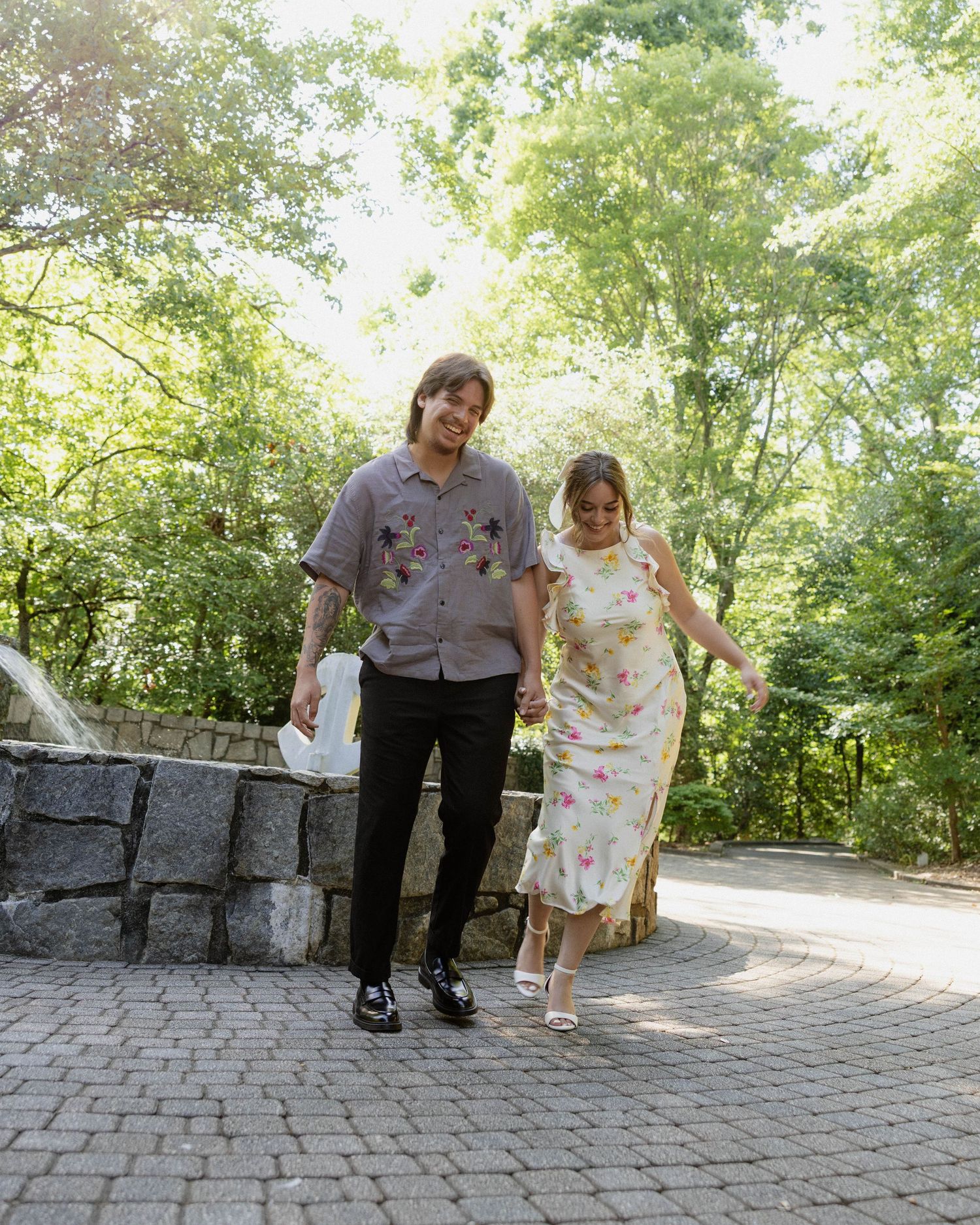 A couple in coordinated floral outfits walks together on a stone pathway surrounded by lush green trees.