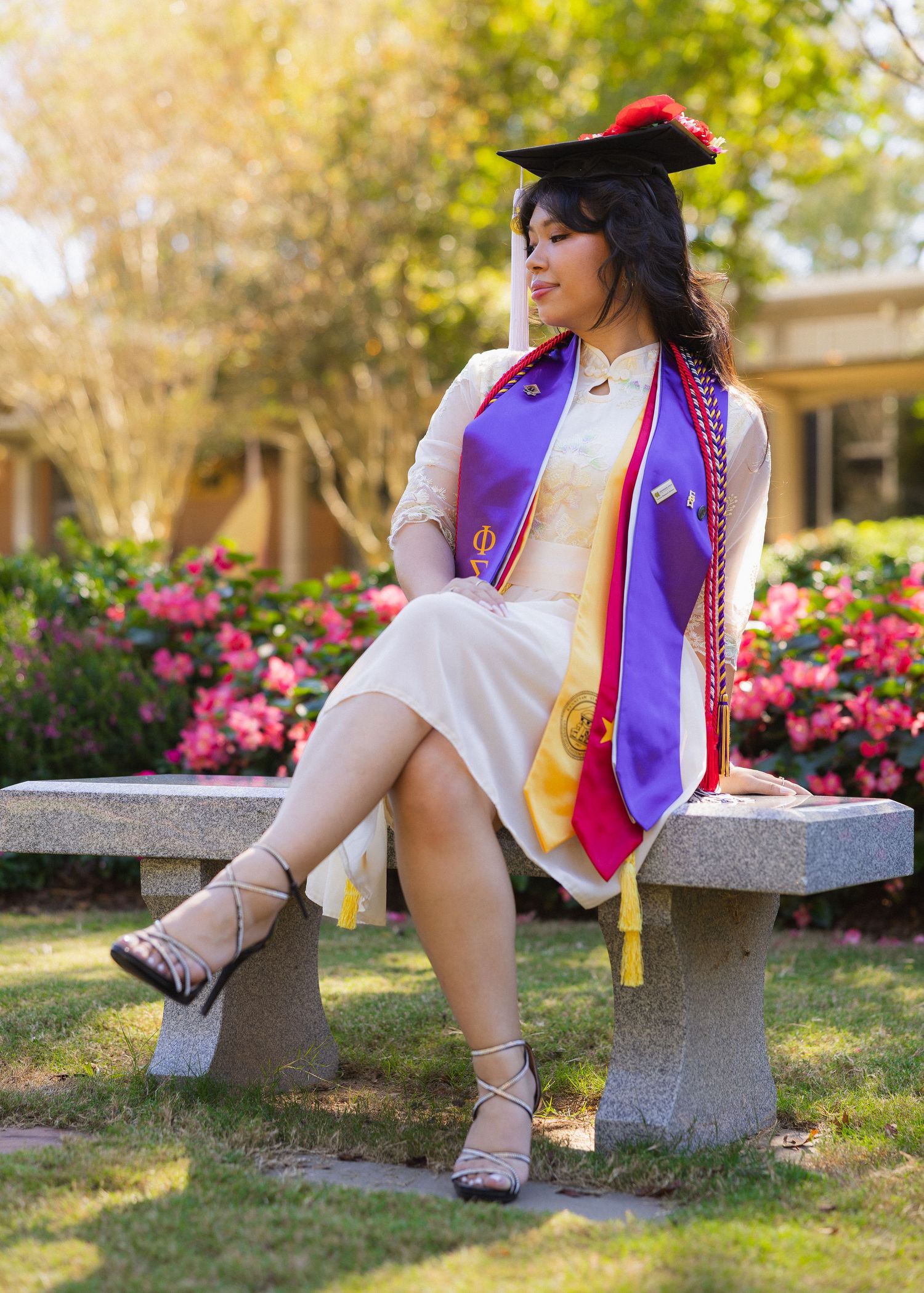 Graduate in purple and white regalia sits on stone bench surrounded by blooming pink flowers in garden setting.