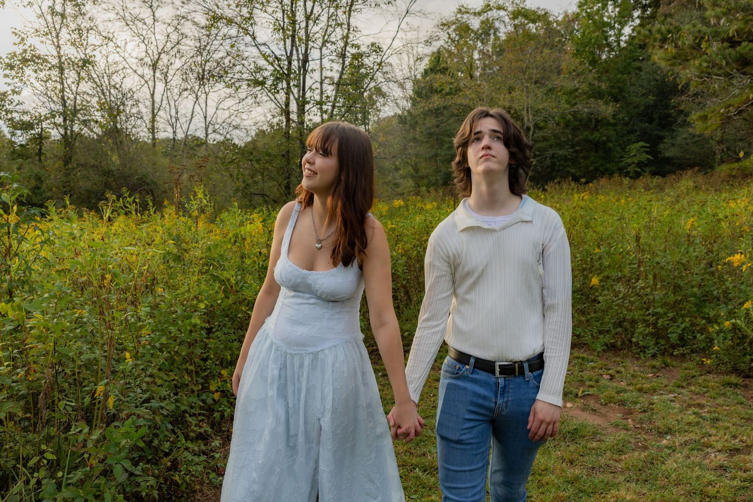 A couple walks hand in hand through a scenic meadow filled with wildflowers and trees on a sunny autumn day.