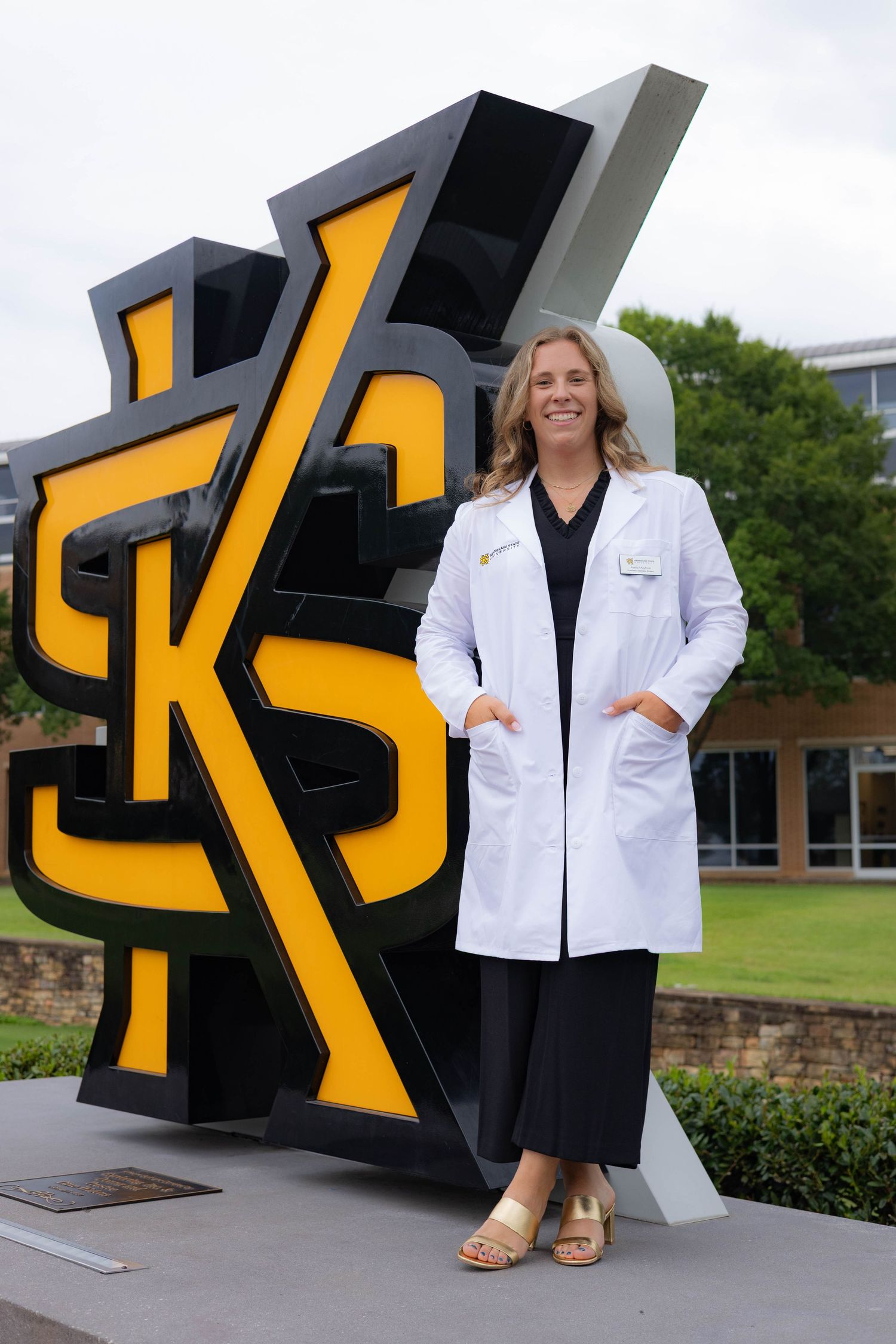 A medical professional in a white coat stands in front of a large black and yellow KSU logo sculpture on campus.
