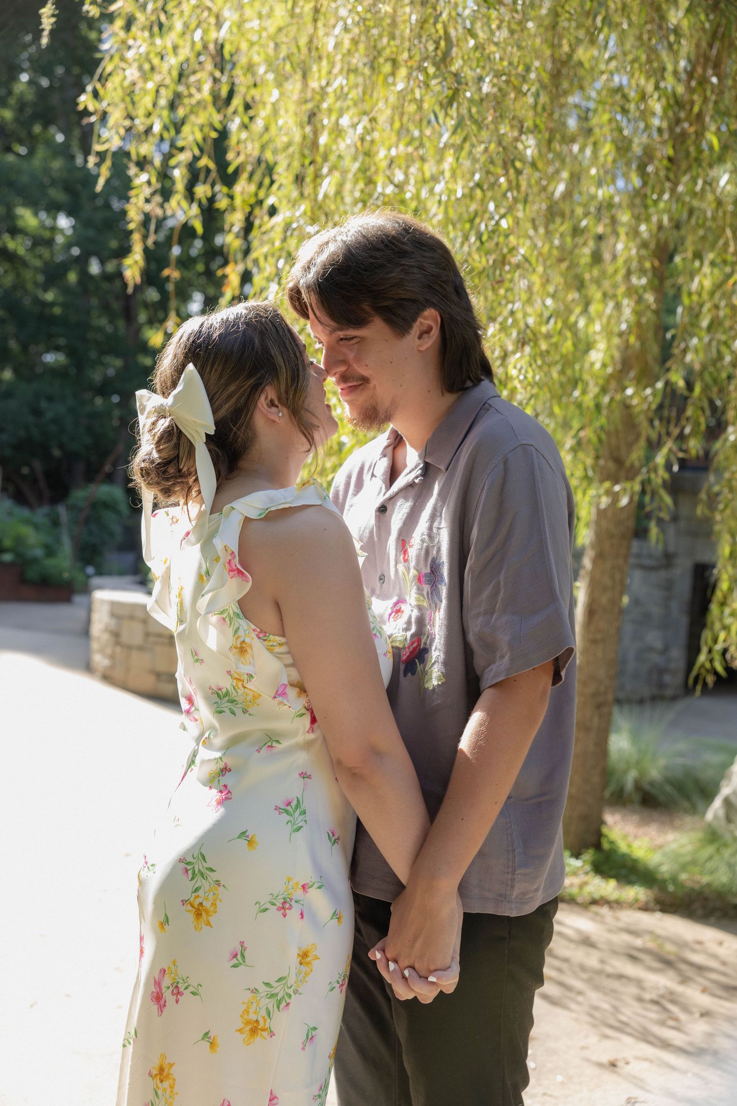 A romantic couple shares a tender embrace under a sunlit willow tree in a dreamy summer setting.