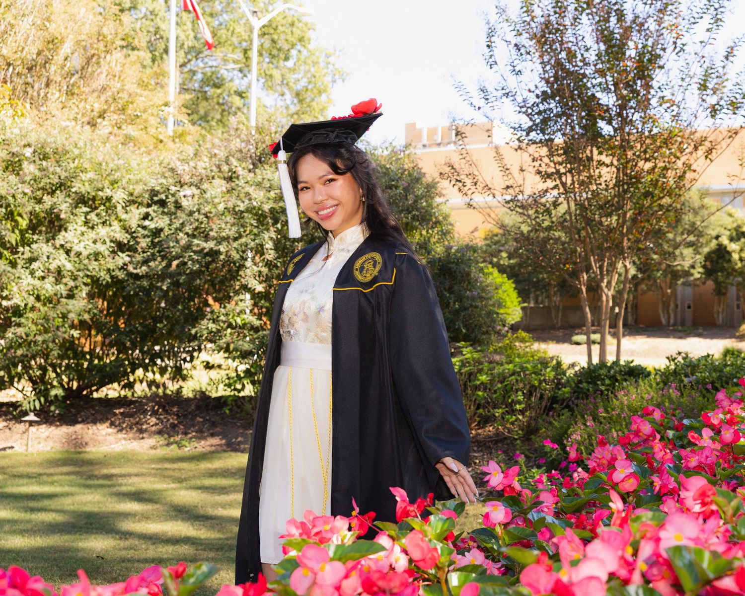 A person in a black jacket poses in a garden with vibrant pink flowers and green foliage in the background.
