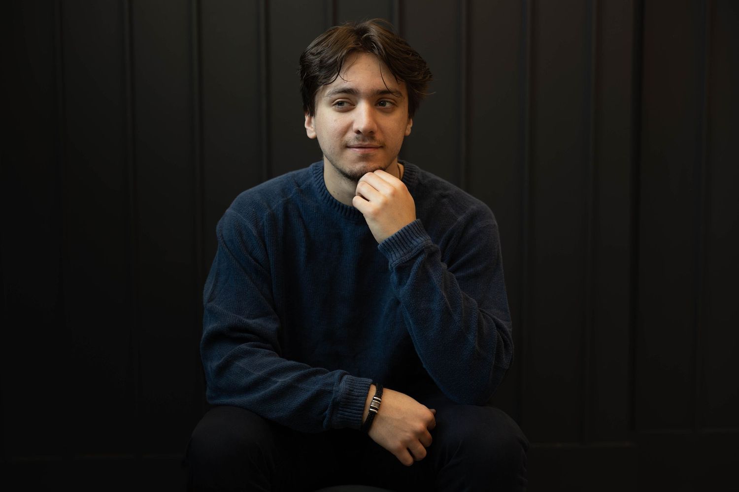 A person in a dark blue sweater poses with a thoughtful expression against a black background.