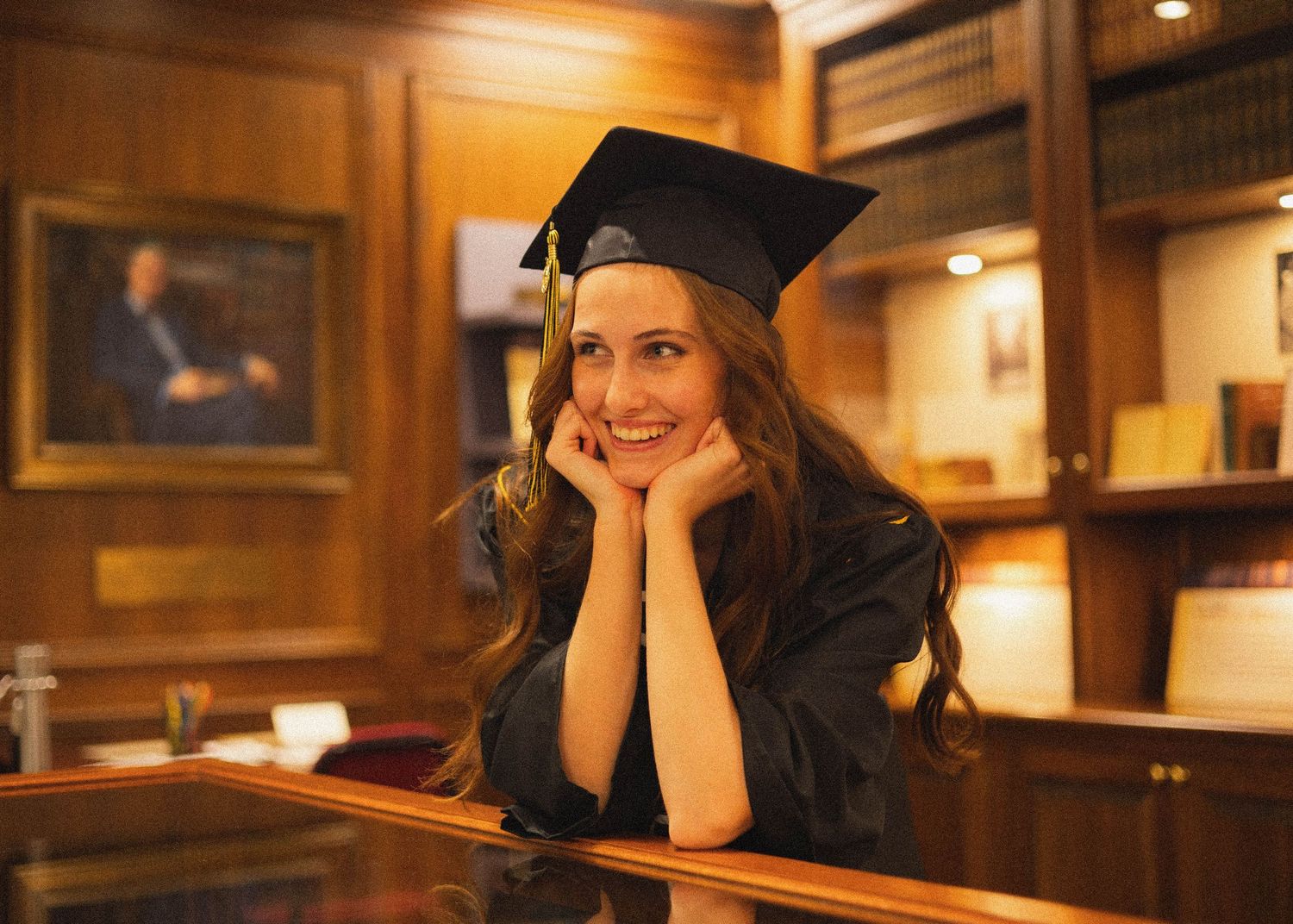 Graduate in cap and gown resting chin on hands in wood-paneled room.