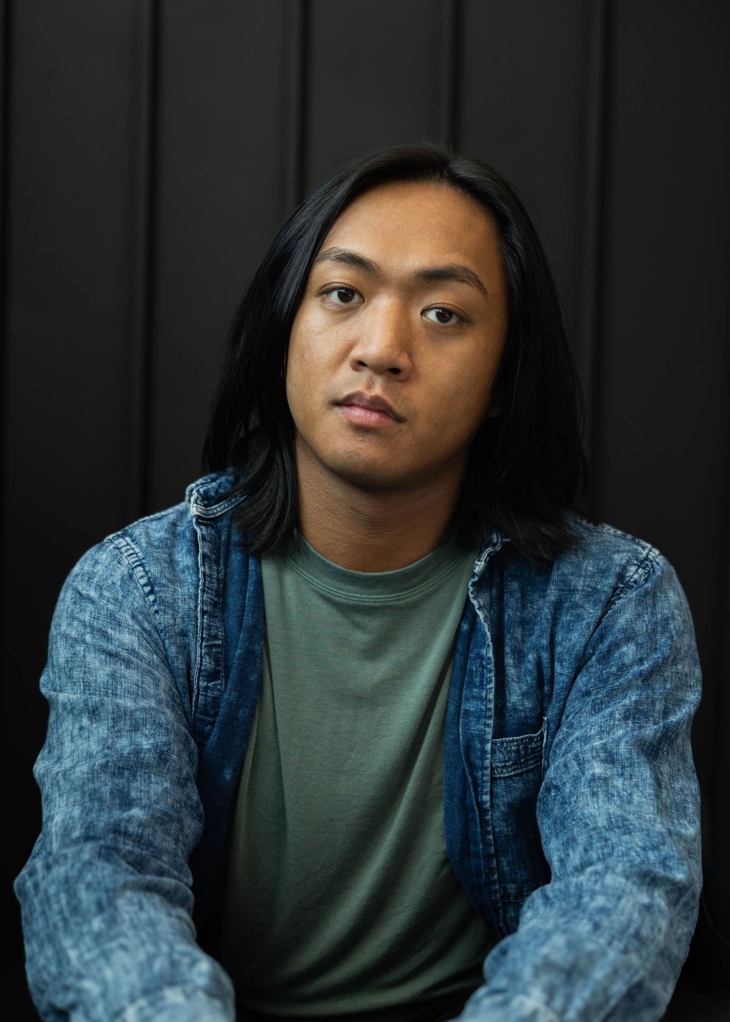 Young person with long black hair wearing a green t-shirt and acid wash denim jacket against dark background.