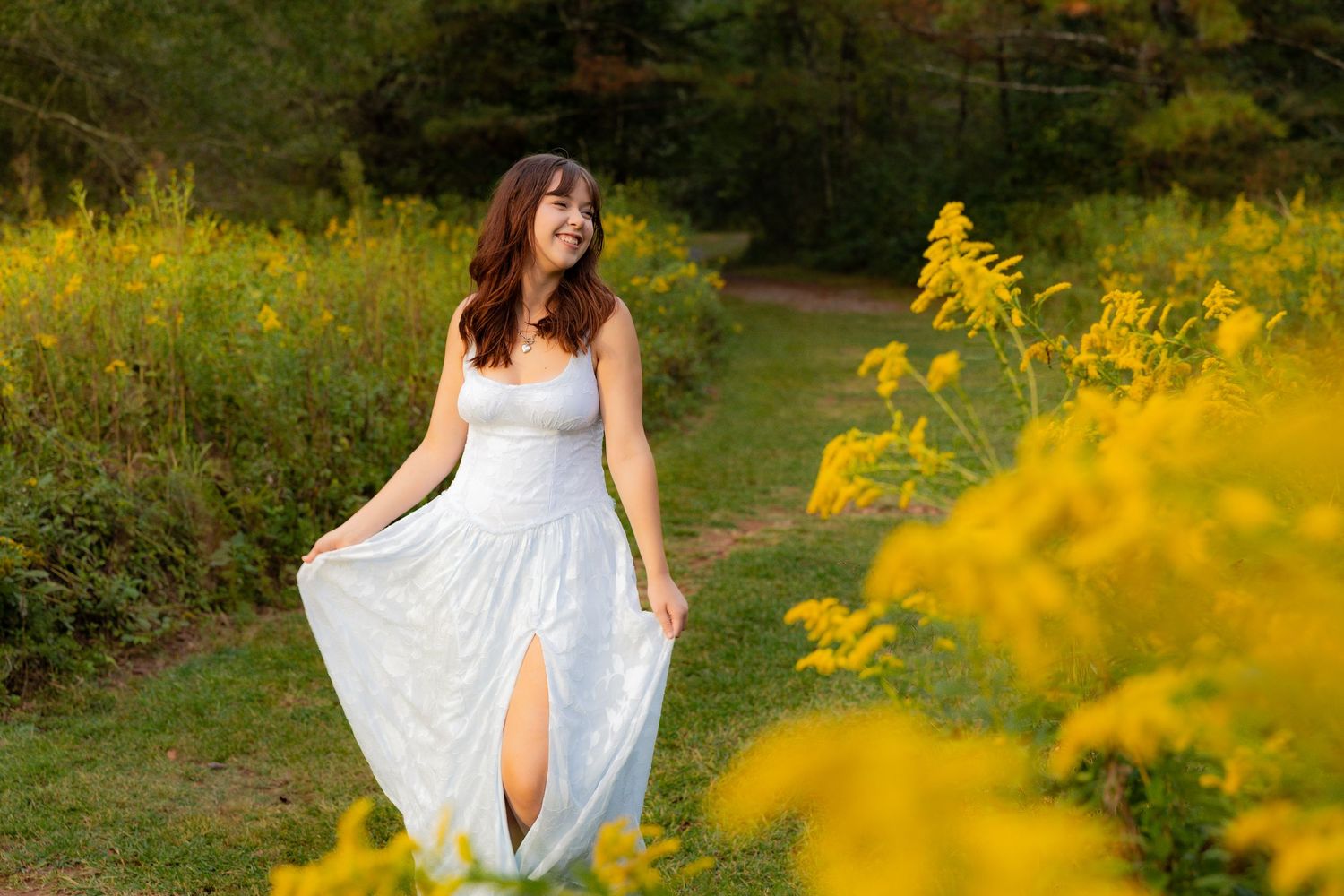 A person in a flowing white dress poses gracefully among vibrant yellow wildflowers in a garden setting.