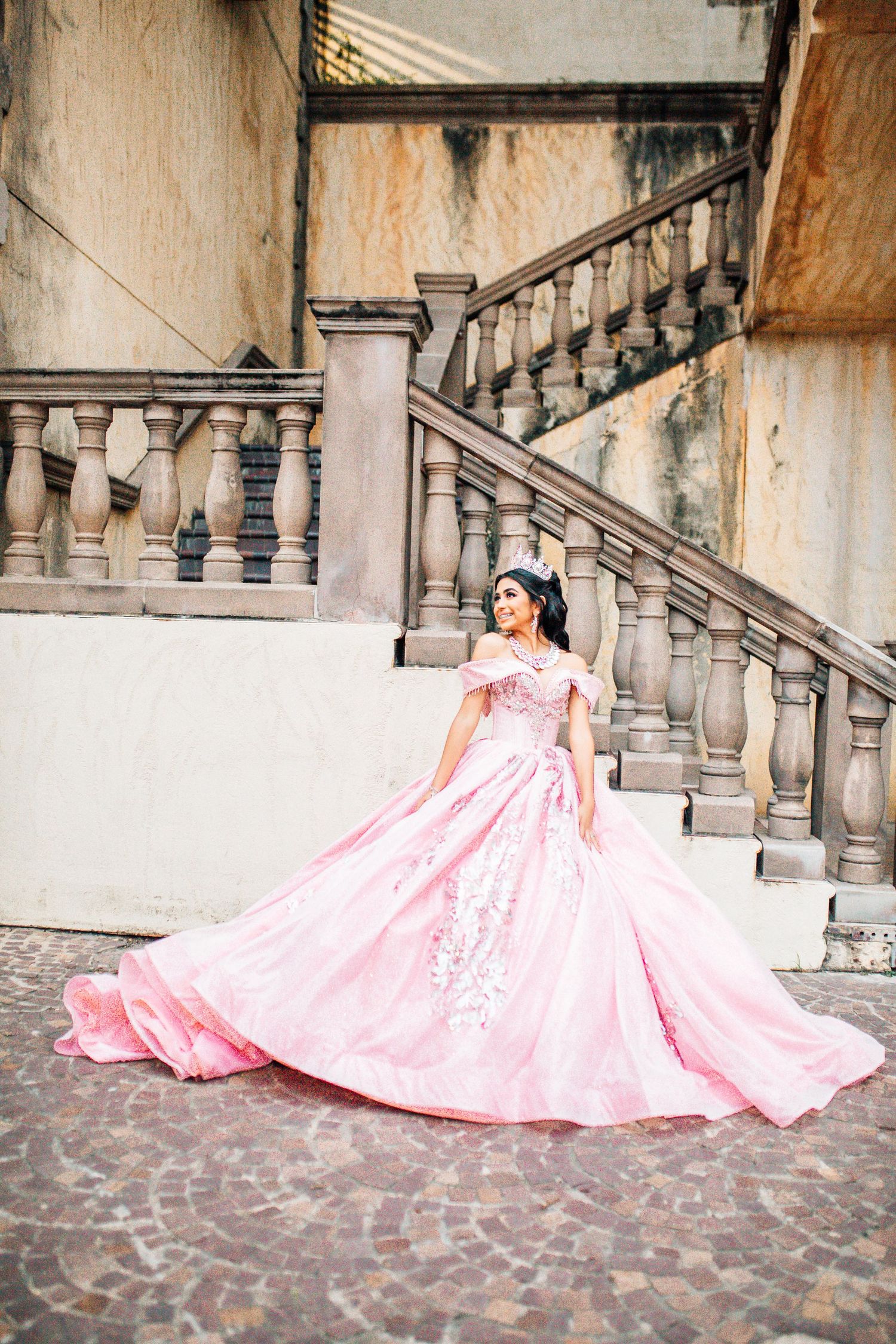 Young woman in a pink ball gown with floral embroidery poses gracefully on cobblestone steps near ornate stone balustrade.