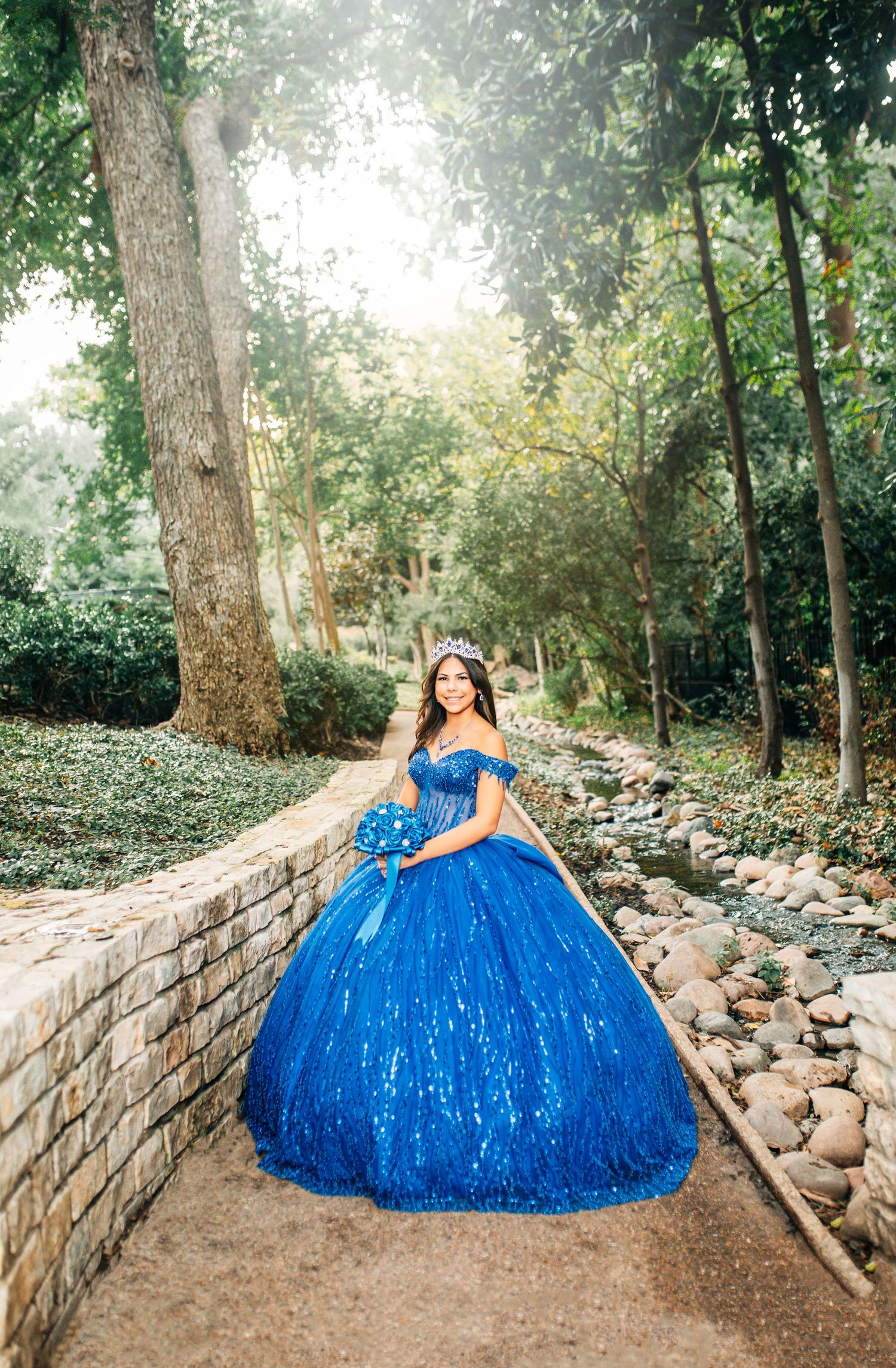 Young woman in a royal blue sequin ball gown and tiara poses in a lush garden for her quinceañera.