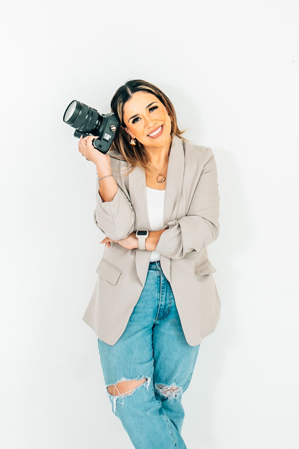 Smiling female photographer holding a DSLR camera, wearing a beige blazer and ripped jeans against a white background.