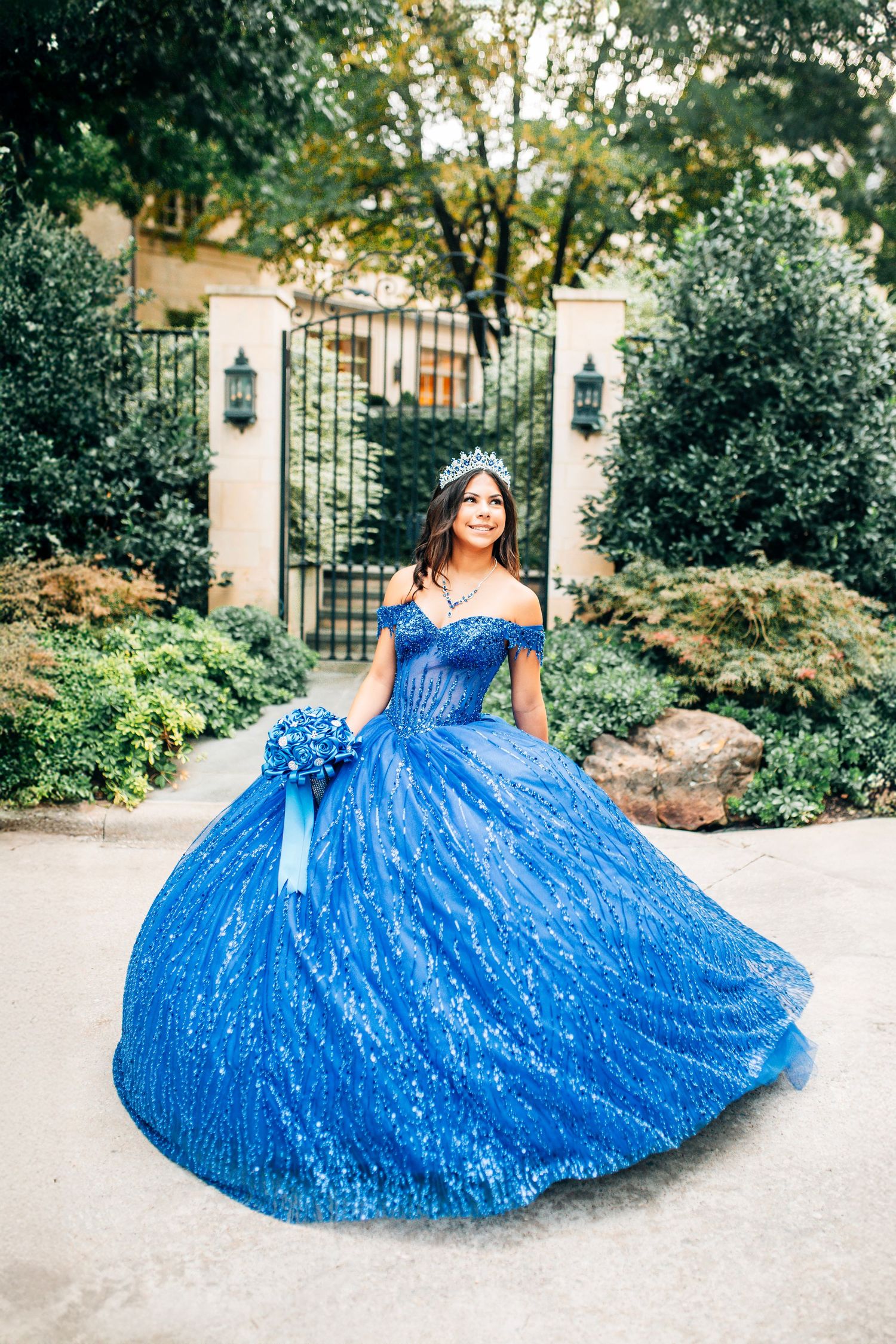 Young woman in a sparkling royal blue quinceañera ball gown with tiara holds bouquet in lush garden.