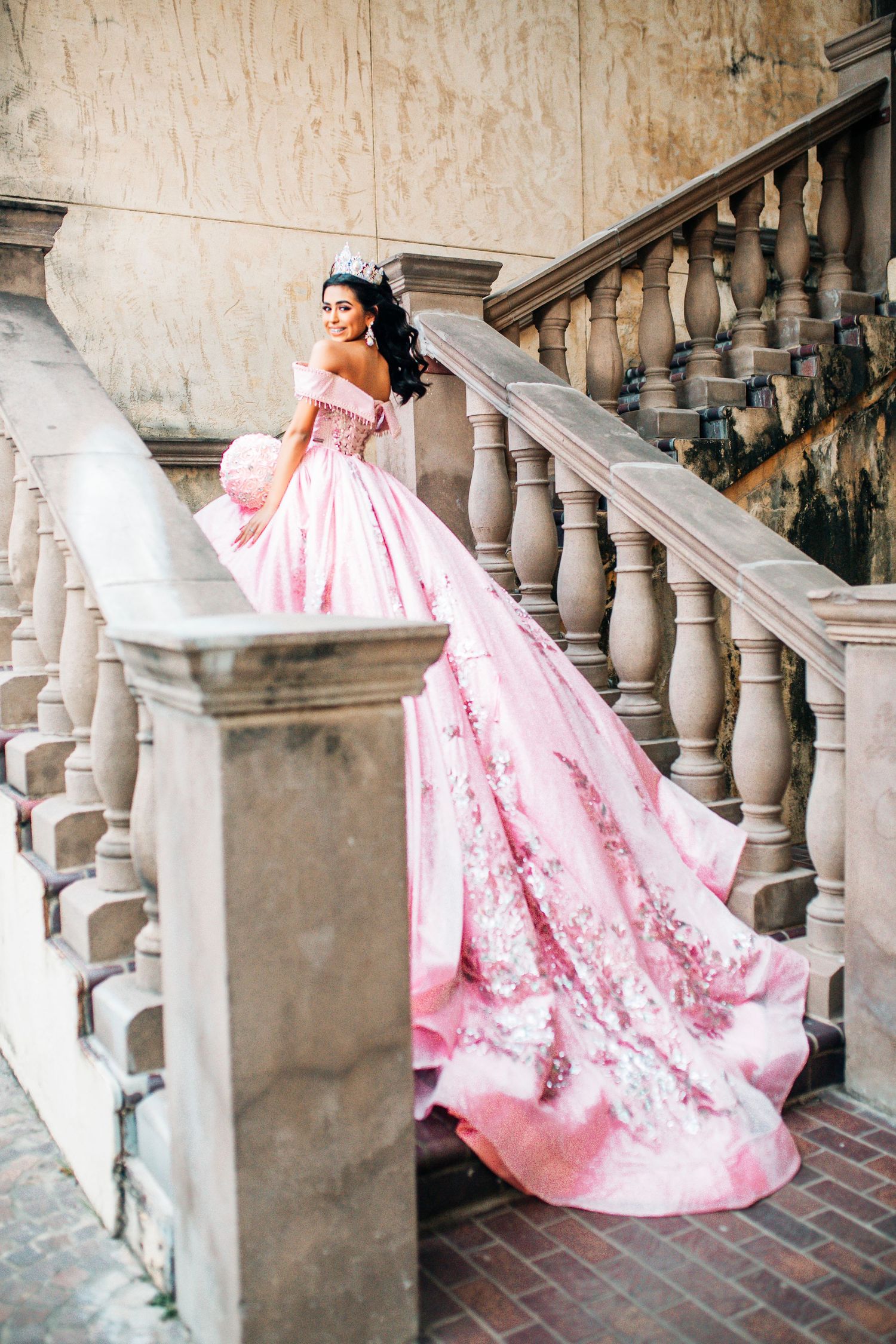Young woman quinceañera in elegant pink ball gown with floral embroidery poses on ornate stone staircase.
