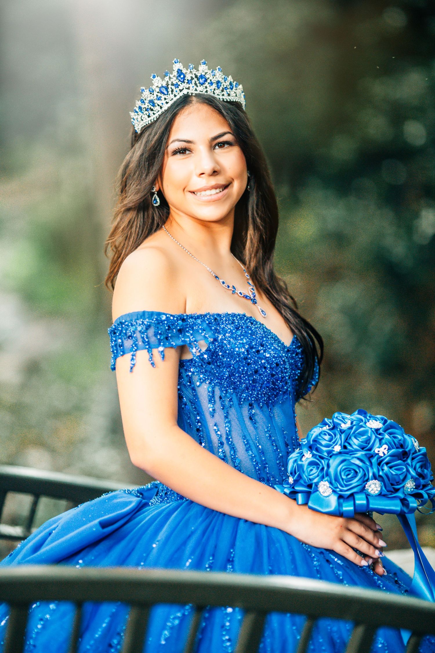 Young woman in blue quinceañera gown with crown and bouquet smiles outdoors.
