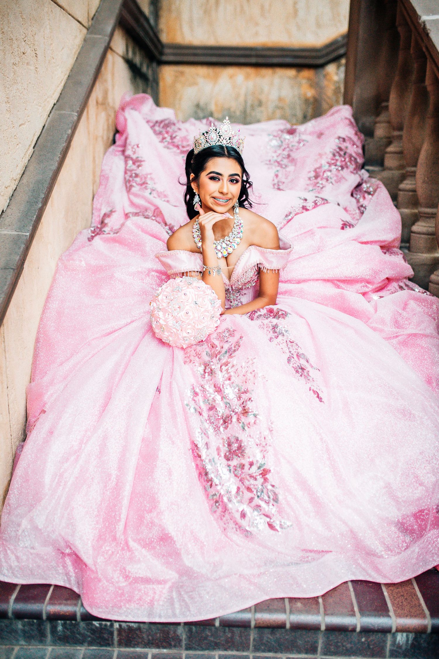 Young woman in a pink quinceañera ball gown with tiara holds bouquet on stone steps.
