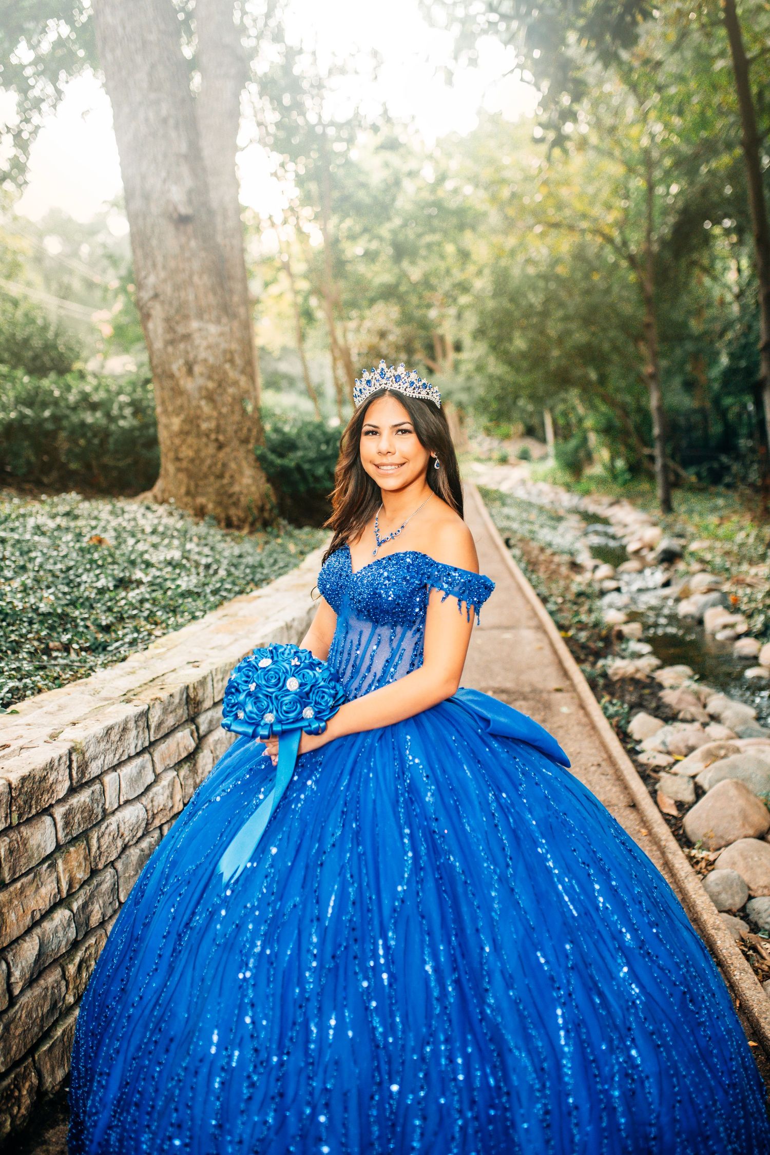 Young woman in a royal blue sequined quinceañera ball gown with tiara and matching bouquet in a garden.