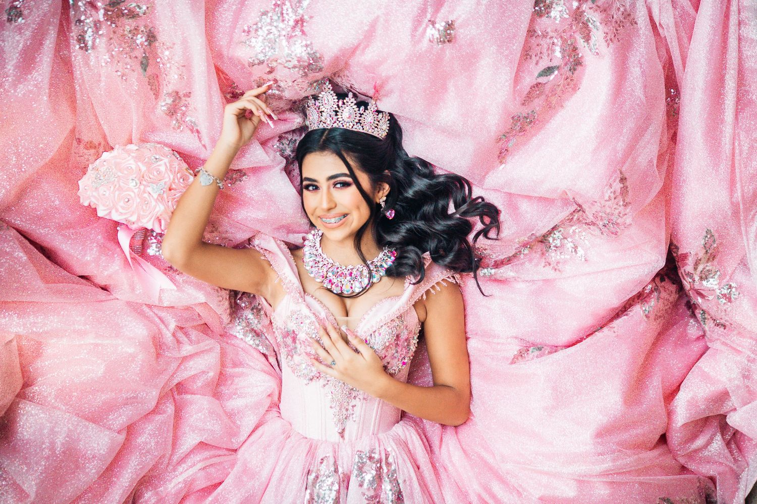 Young woman in pink ballgown and crystal crown poses elegantly among flowing pink fabric for quinceañera portrait.
