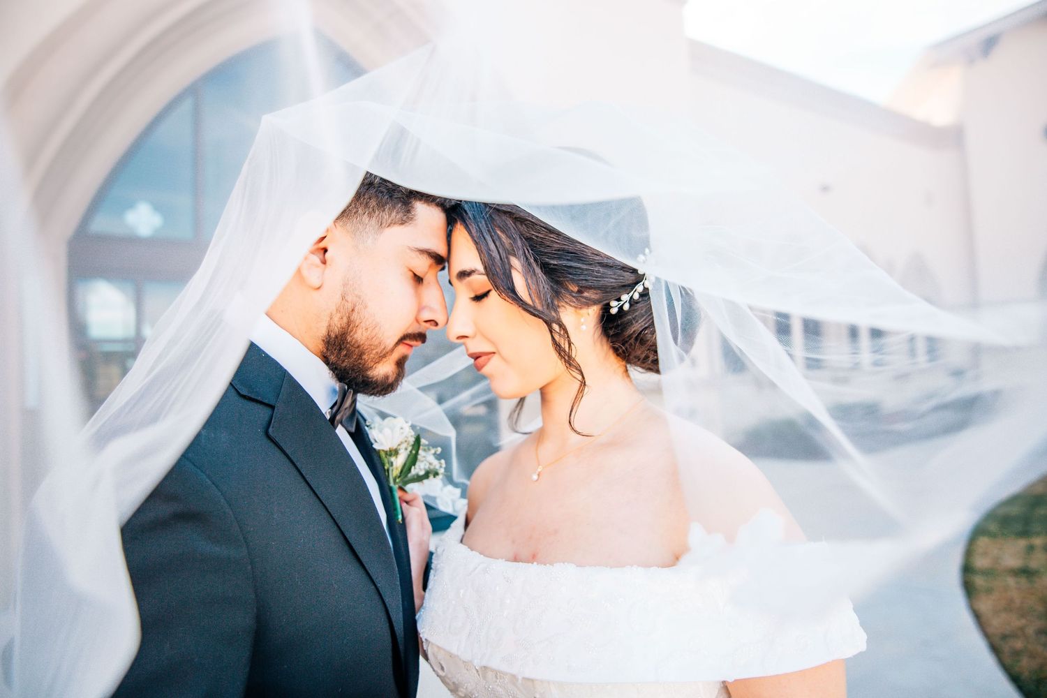 Bride and groom share an intimate moment under a flowing white veil outdoors on their wedding day.