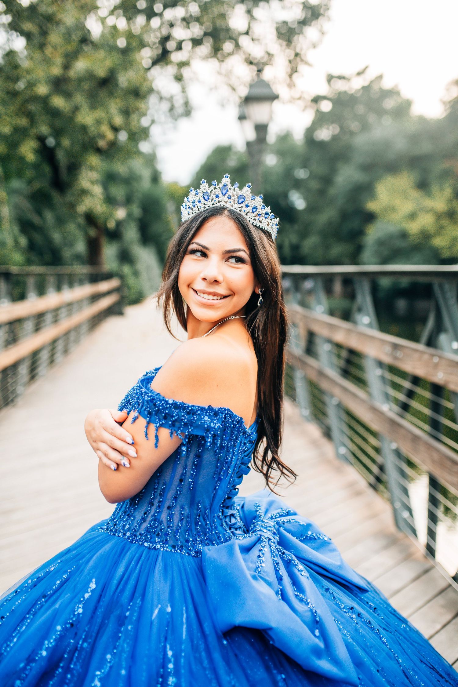 Young woman in blue quinceañera gown and crystal crown smiles on wooden bridge surrounded by lush greenery.