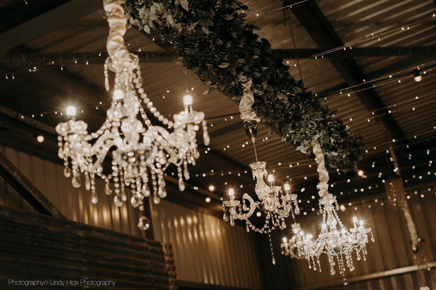 Three elegant crystal chandeliers hang from a rustic wooden ceiling adorned with string lights and dark foliage.