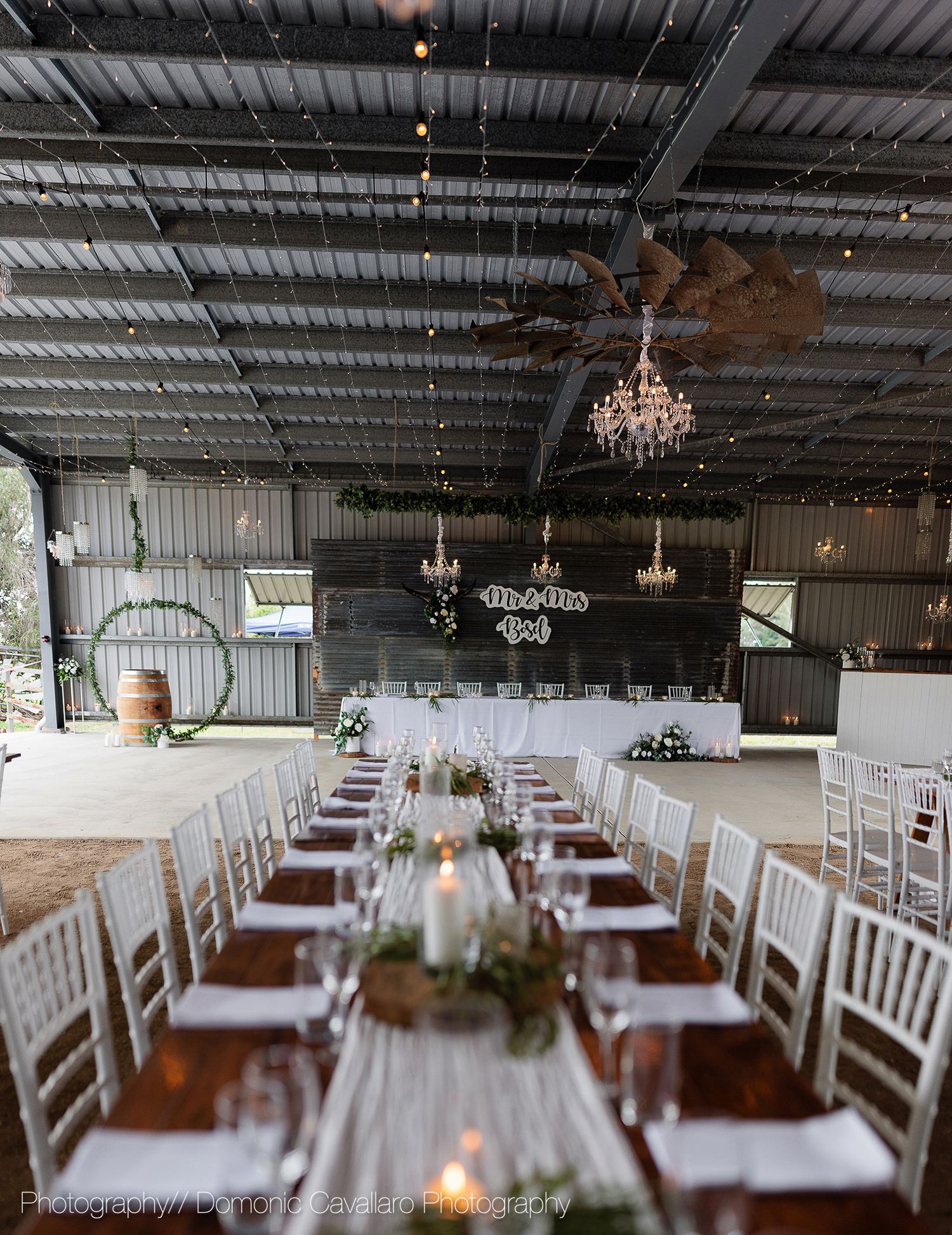Wedding reception venue with white Chiavari chairs and wooden tables arranged under industrial ceiling with chandeliers.