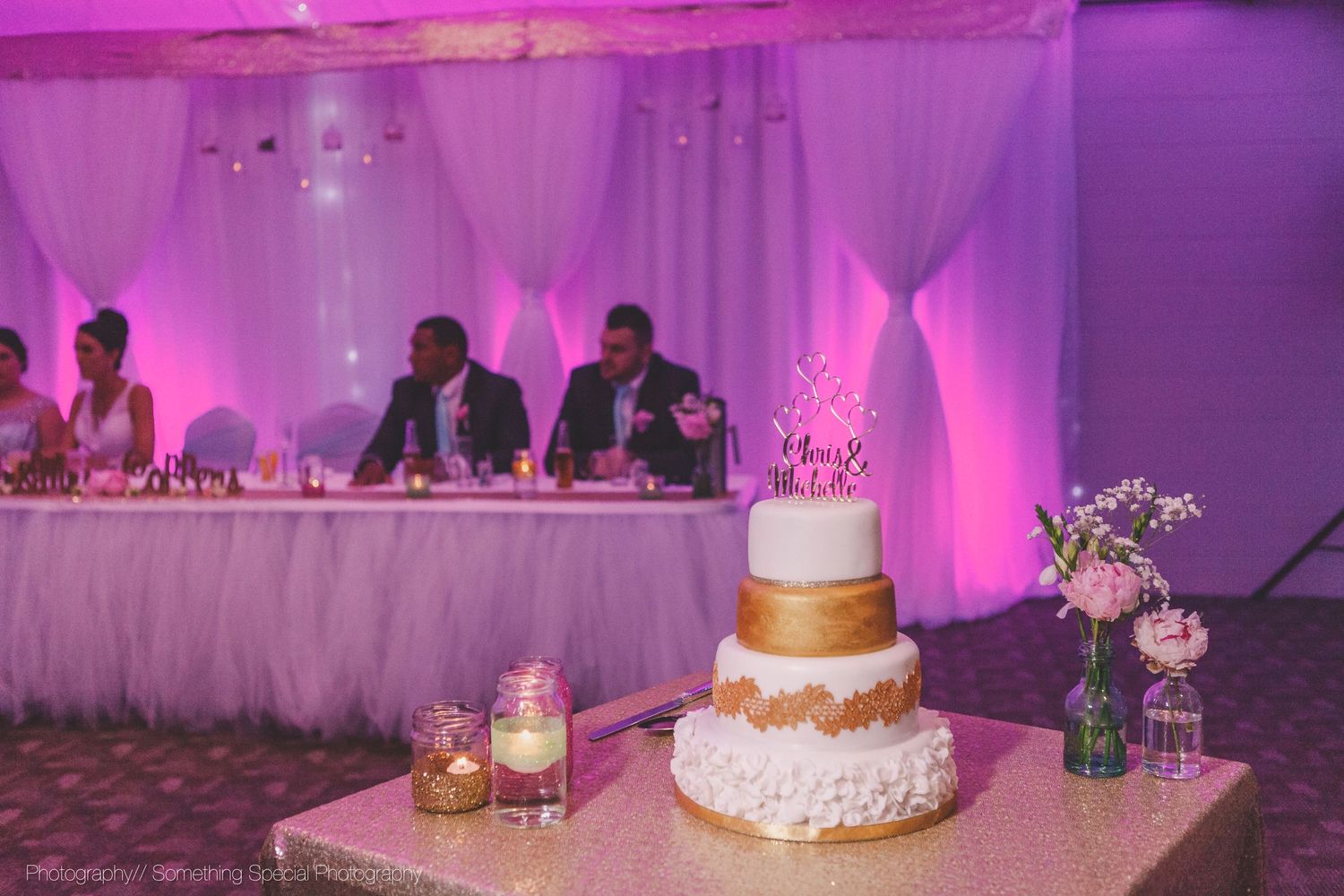 A three-tier white and gold wedding cake displayed at reception with purple uplighting and head table in background.