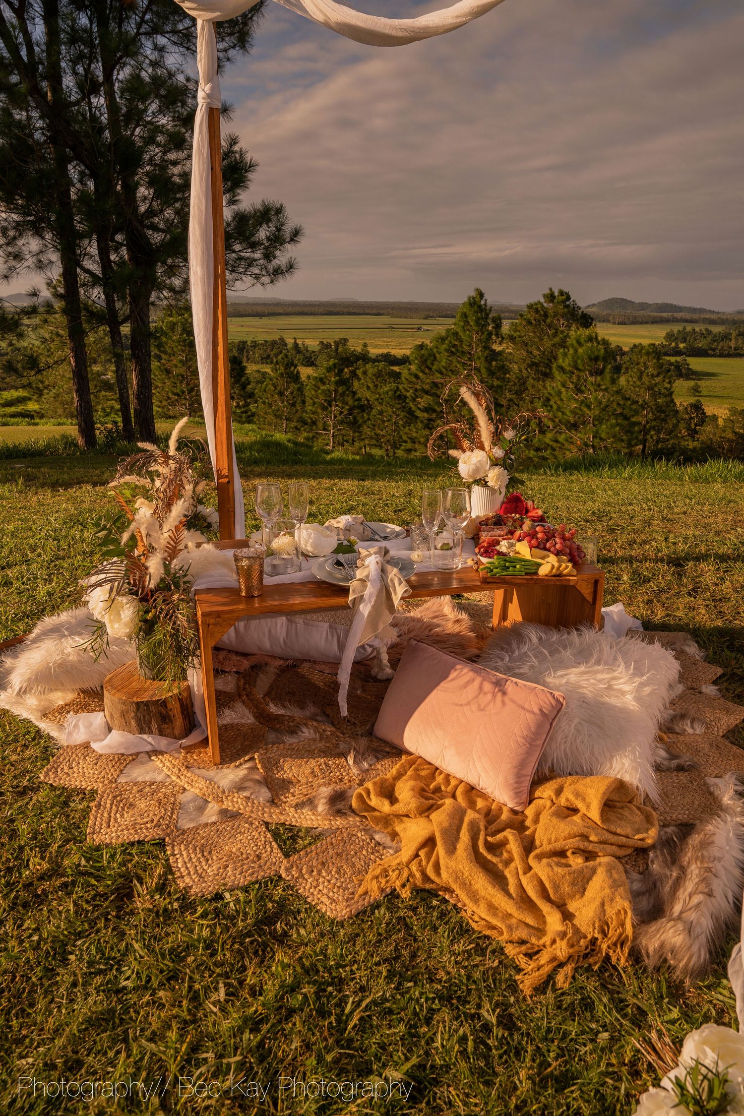A cozy outdoor picnic setup with plush pillows, blankets and decor overlooking a scenic hillside landscape at sunset.