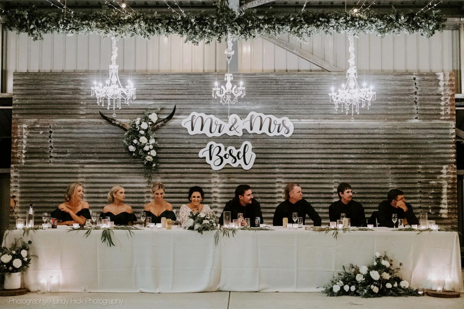 Wedding reception venue with elegant crystal chandeliers and bridal party seated at long table under Mr and Mrs text decor.