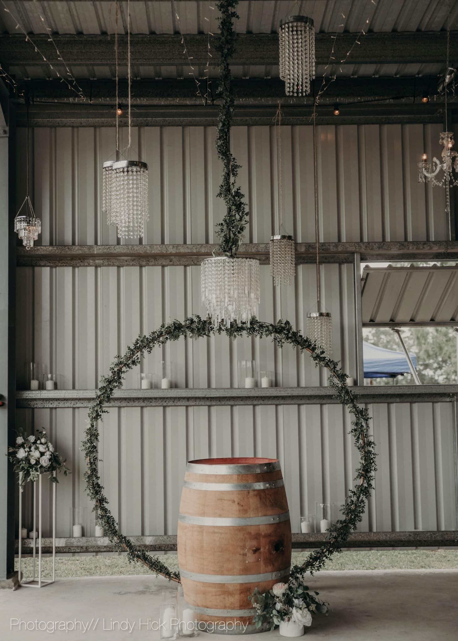 Rustic barn wedding decor with crystal chandeliers hanging above a wooden barrel surrounded by greenery hoops.