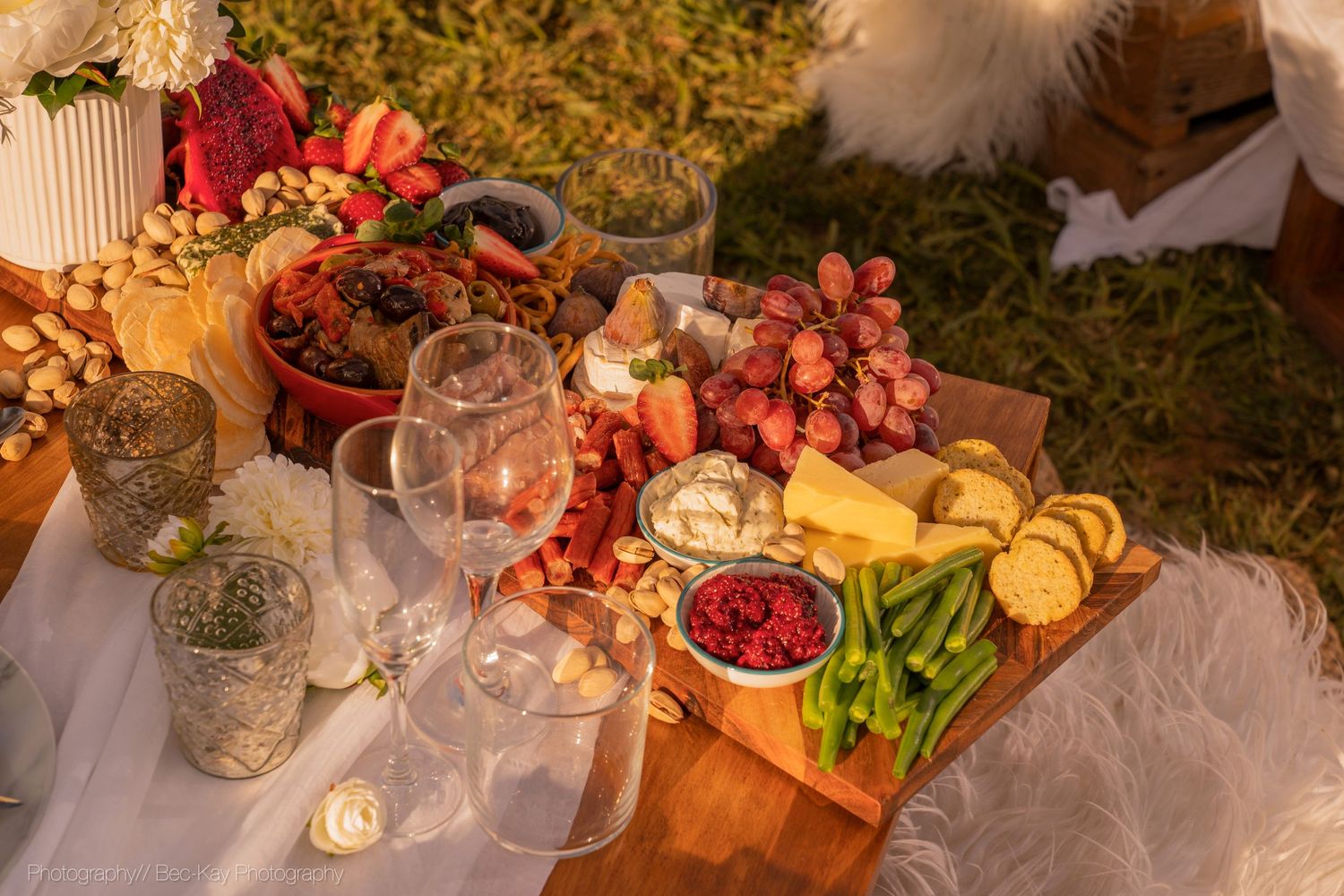 Rustic wooden charcuterie board with fresh fruits, cheese, wine glasses and snacks at an outdoor picnic setting.