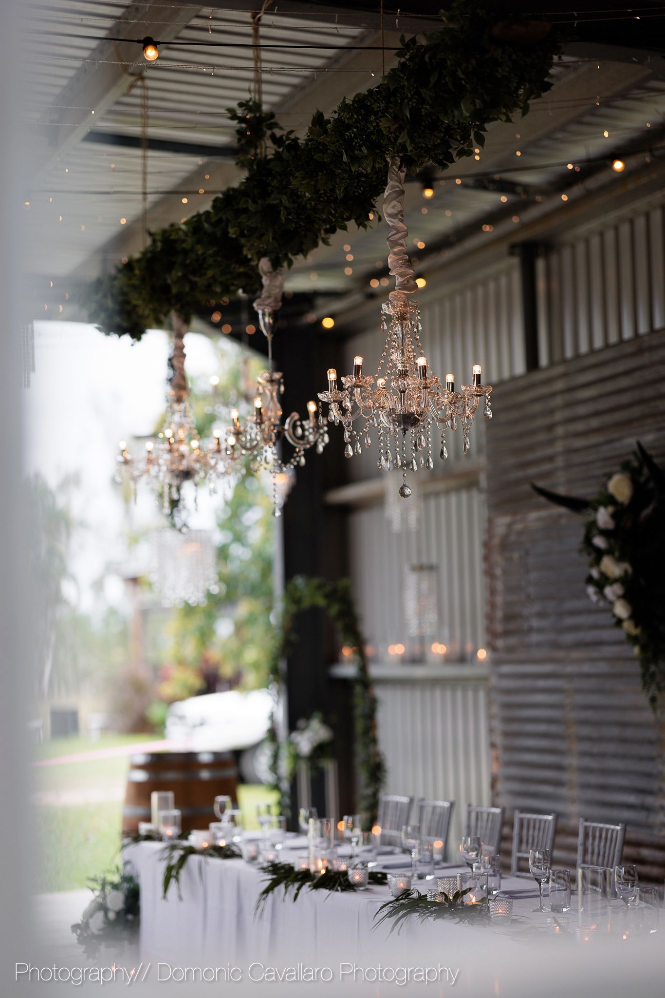 Elegant crystal chandelier and greenery garland create romantic ambiance in rustic barn venue.