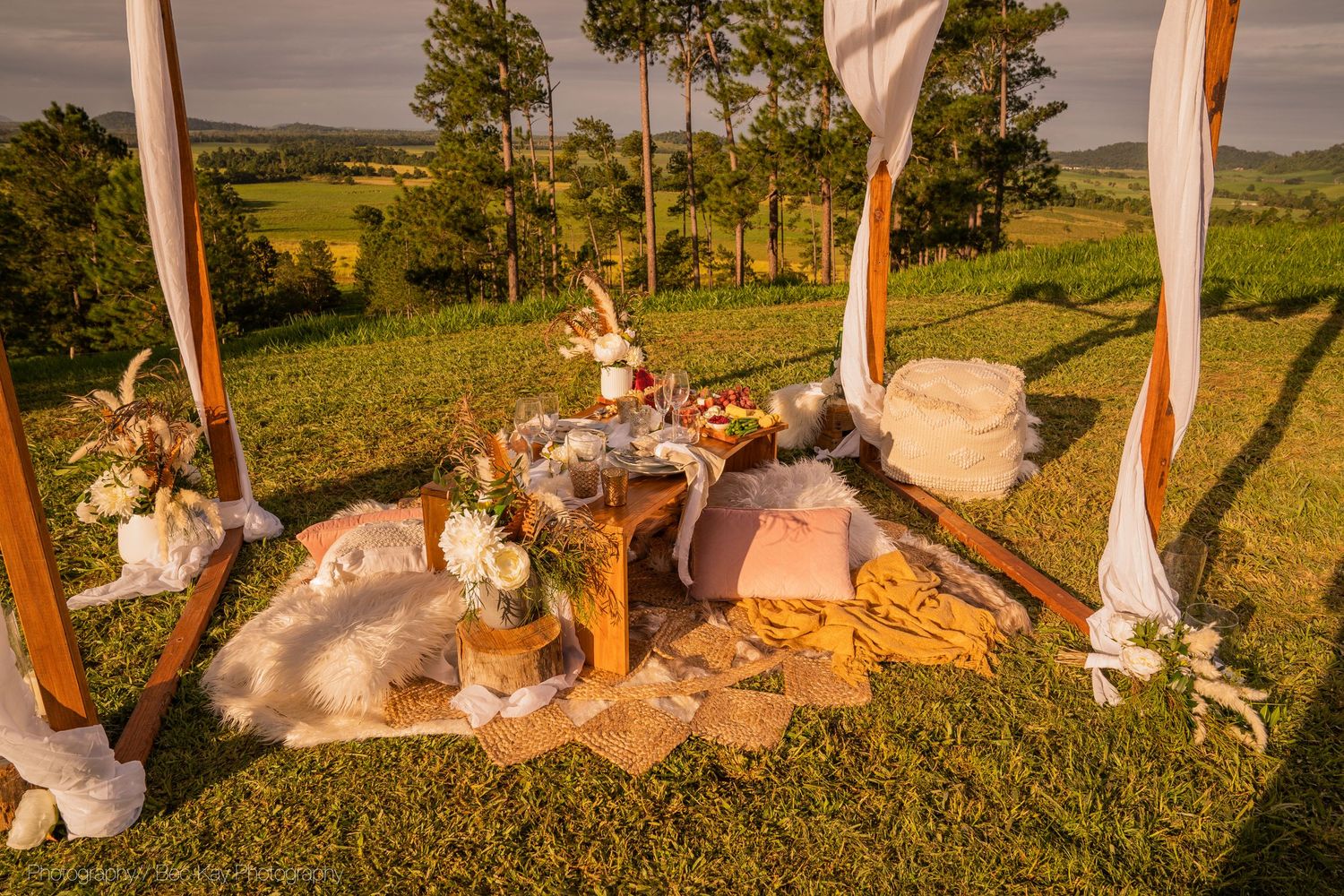 Rustic outdoor picnic setup with plush cushions, pillows and fabric drapes on a grassy hillside at sunset.