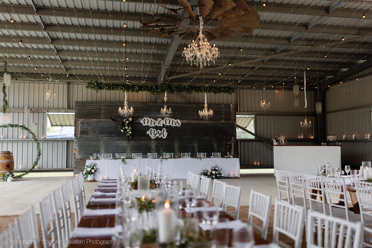 Industrial barn wedding reception venue with crystal chandeliers and white chiavari chairs arranged at dining tables.