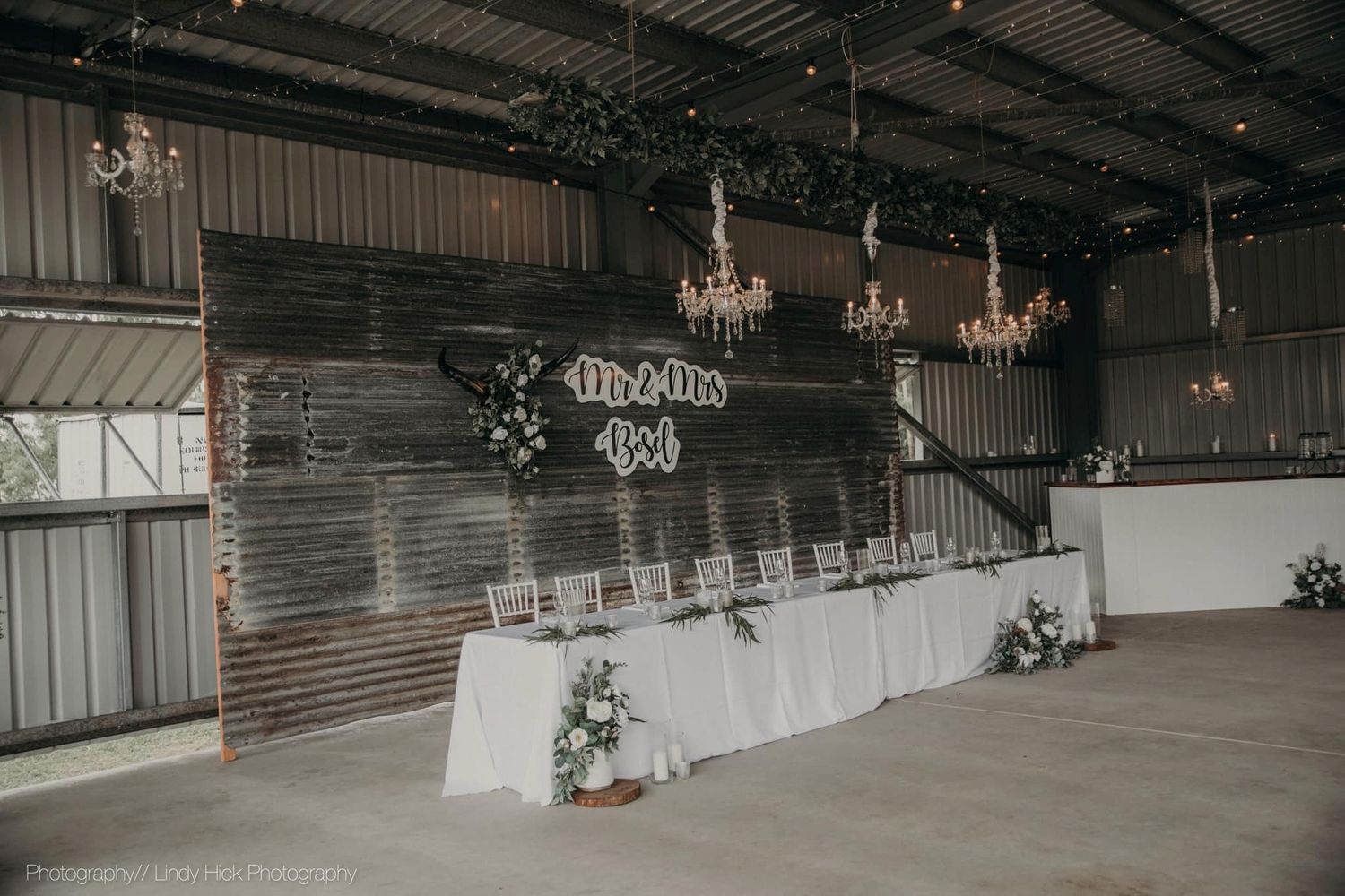 Wedding reception venue with rustic barn walls, chandeliers, and a long white head table decorated with floral arrangements.